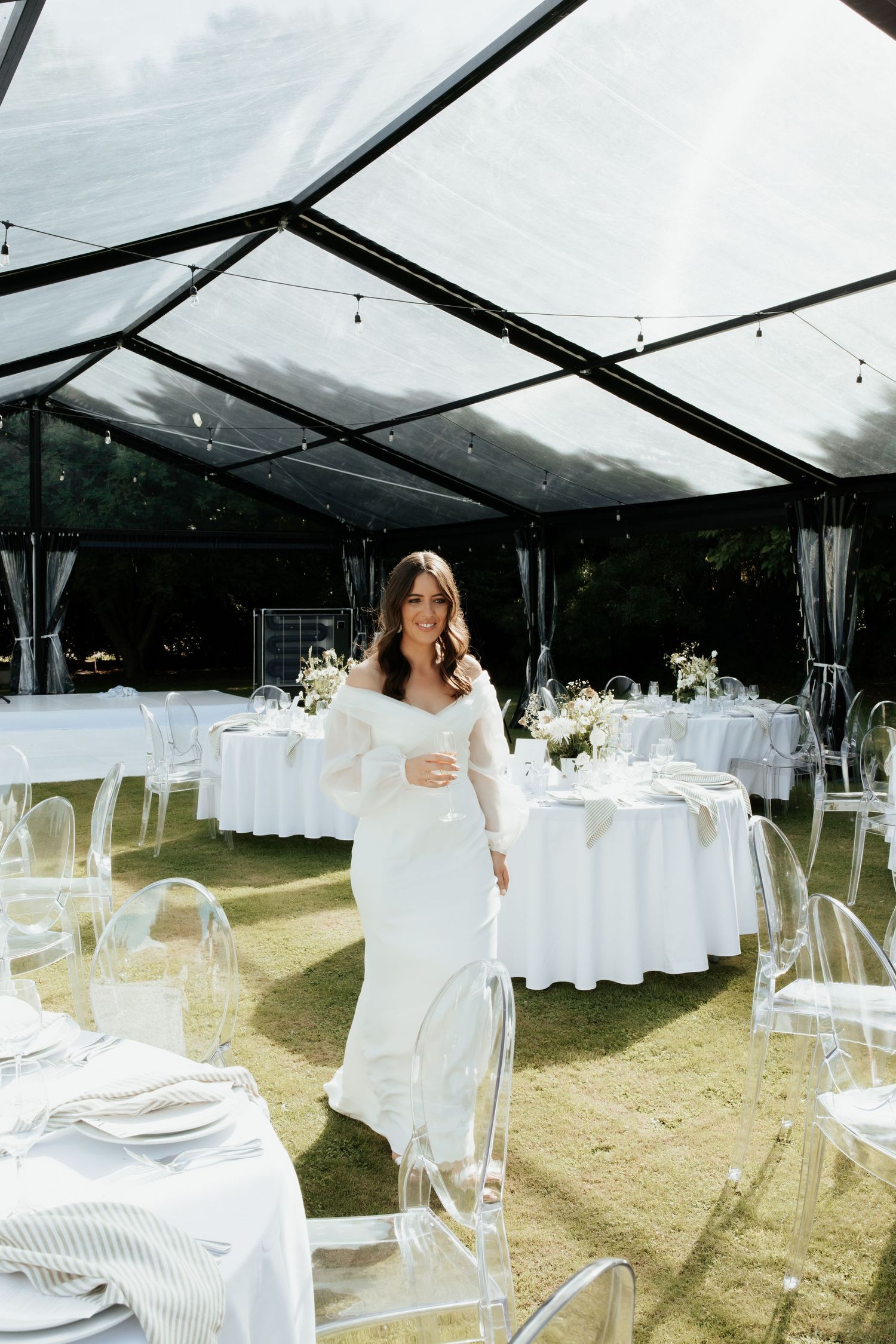 Modern wedding reception setup under clear tent with ghost chairs and white tablecloths.
