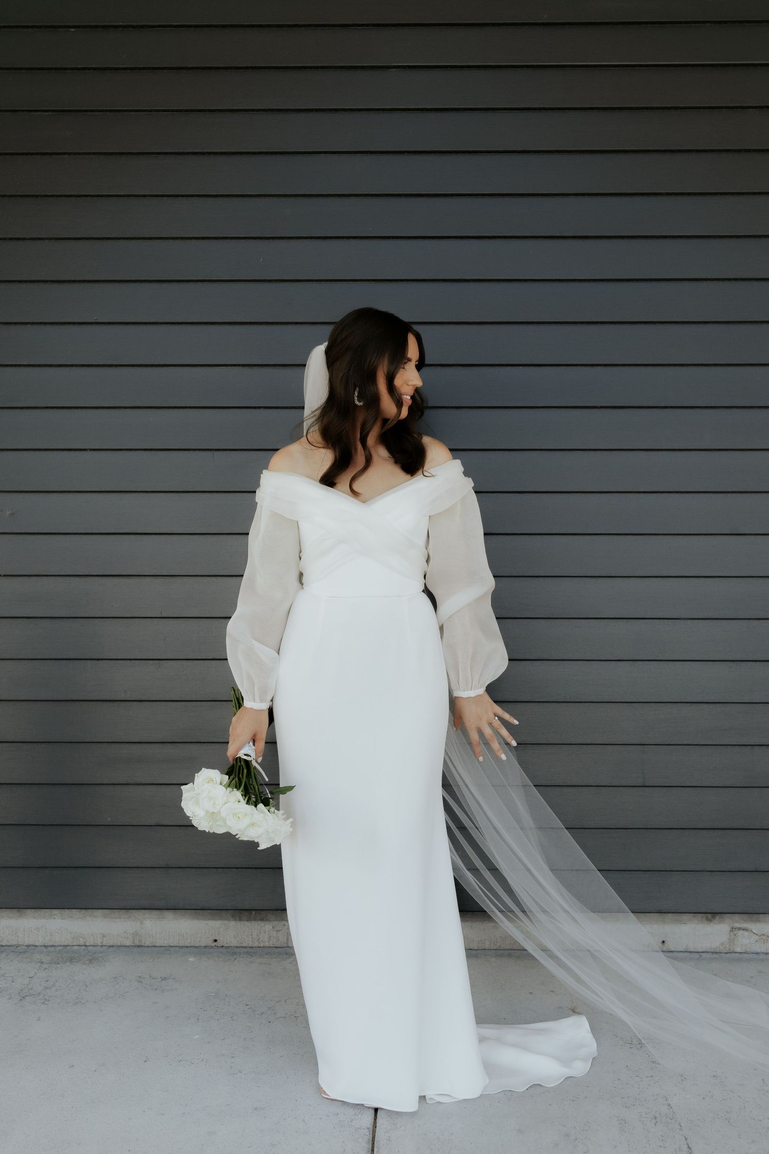 Side view of bride in off-shoulder gown with flowing veil against gray wall.