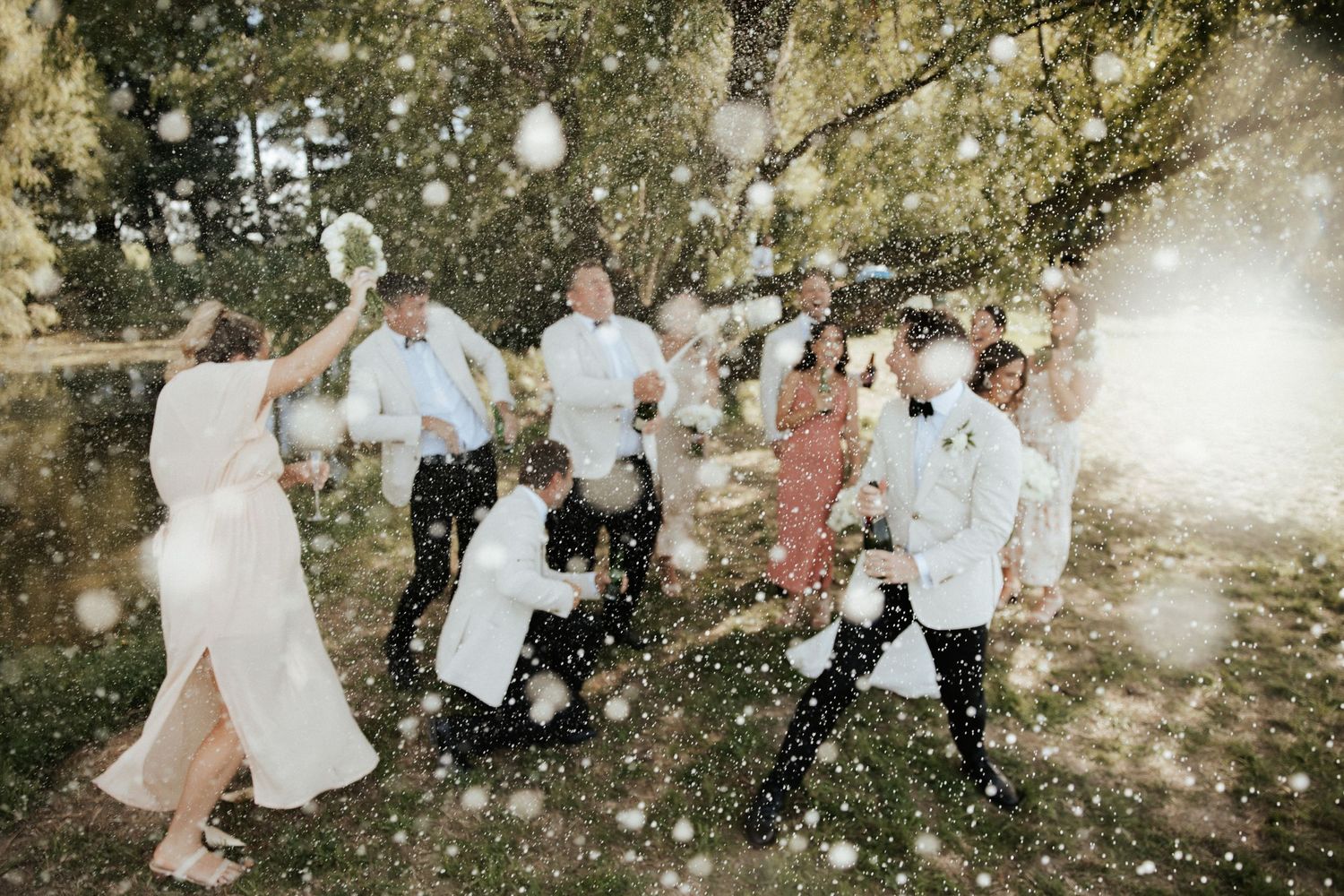 Wedding party dances and celebrates amid floating confetti in a sunlit garden setting.
