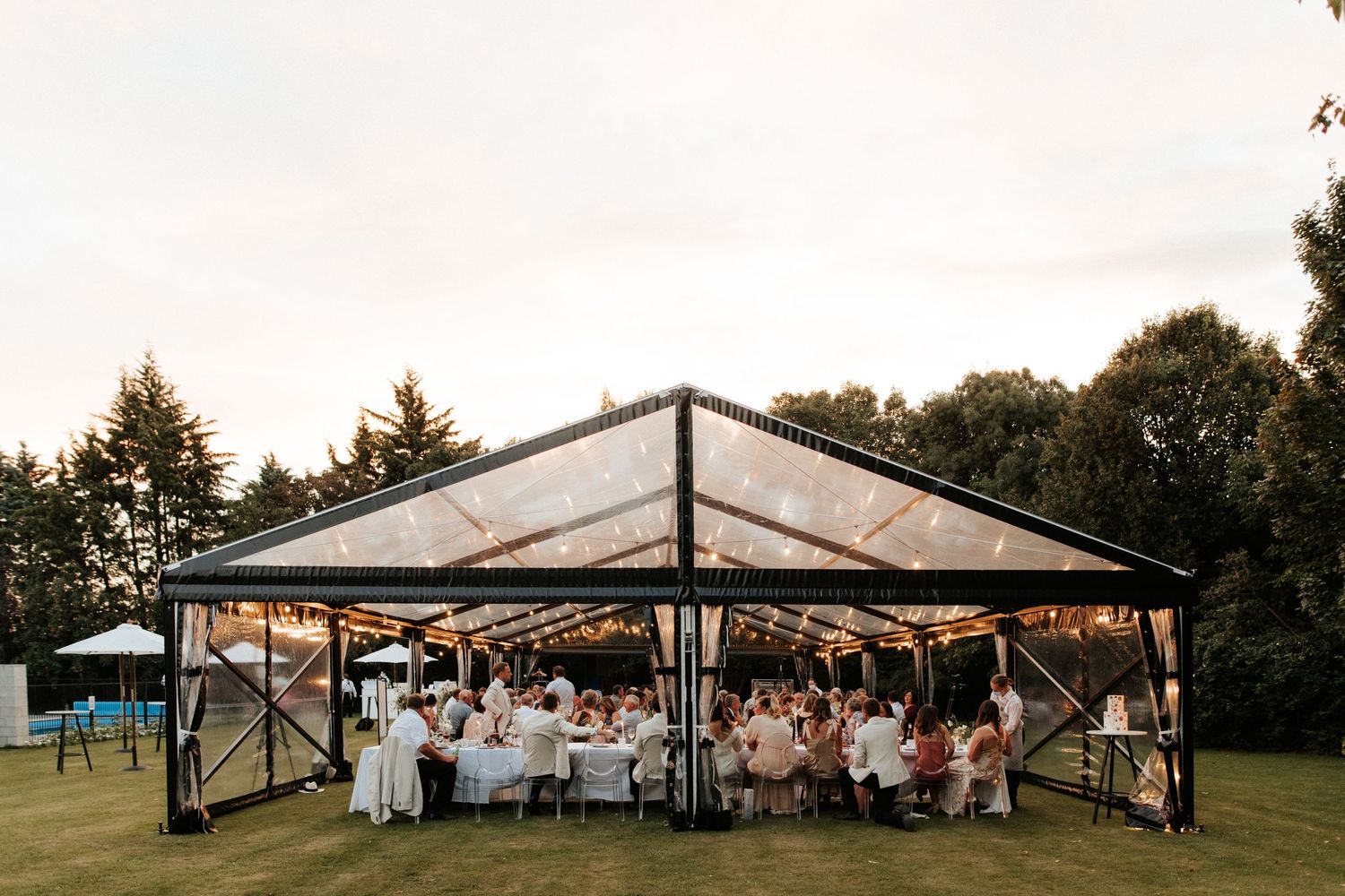 Wedding reception setup under a clear tent pavilion with guests seated at decorated tables.