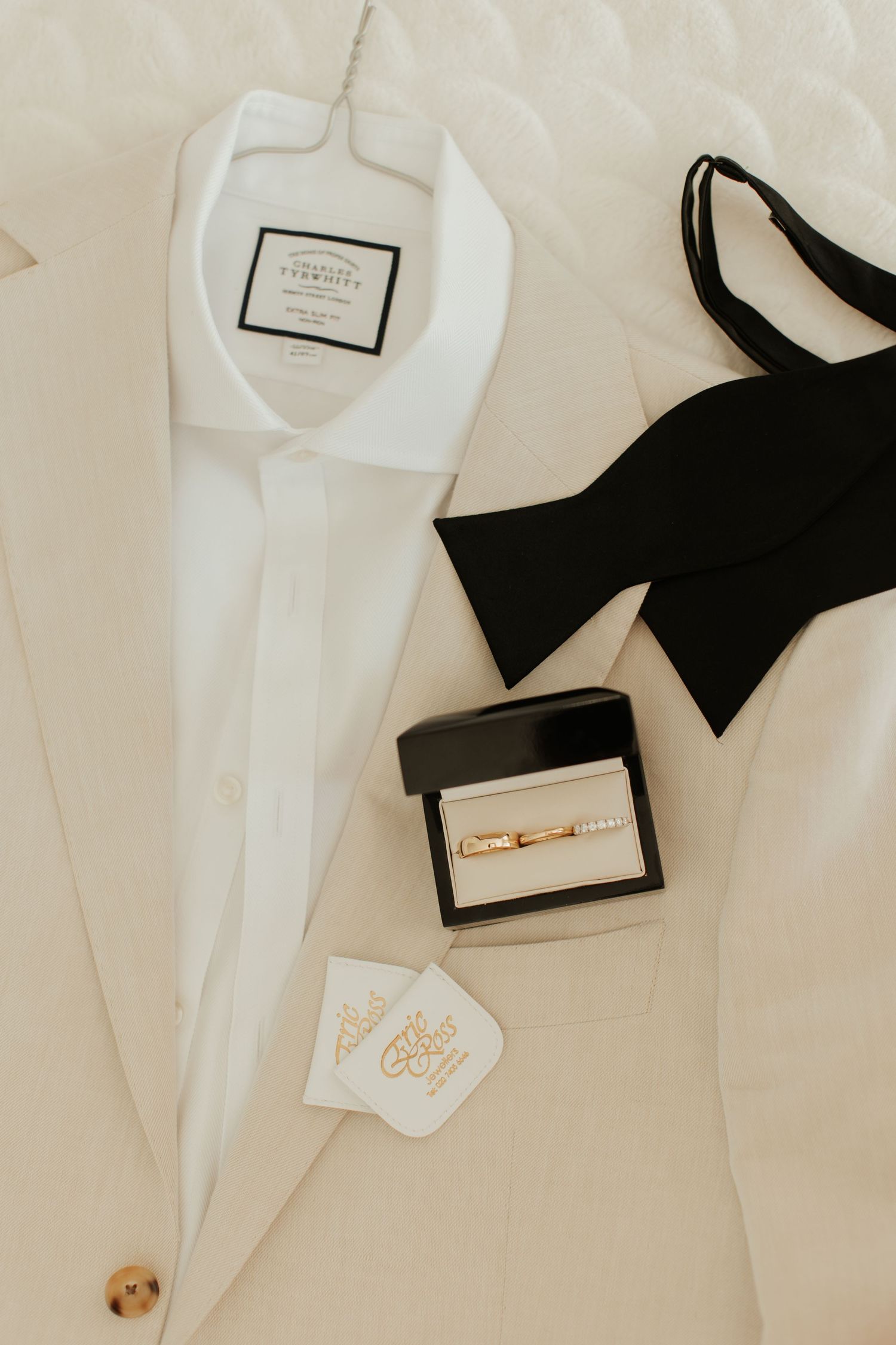 Wedding details flatlay with white dress shirt, black bow tie, and ring box on quilted surface.