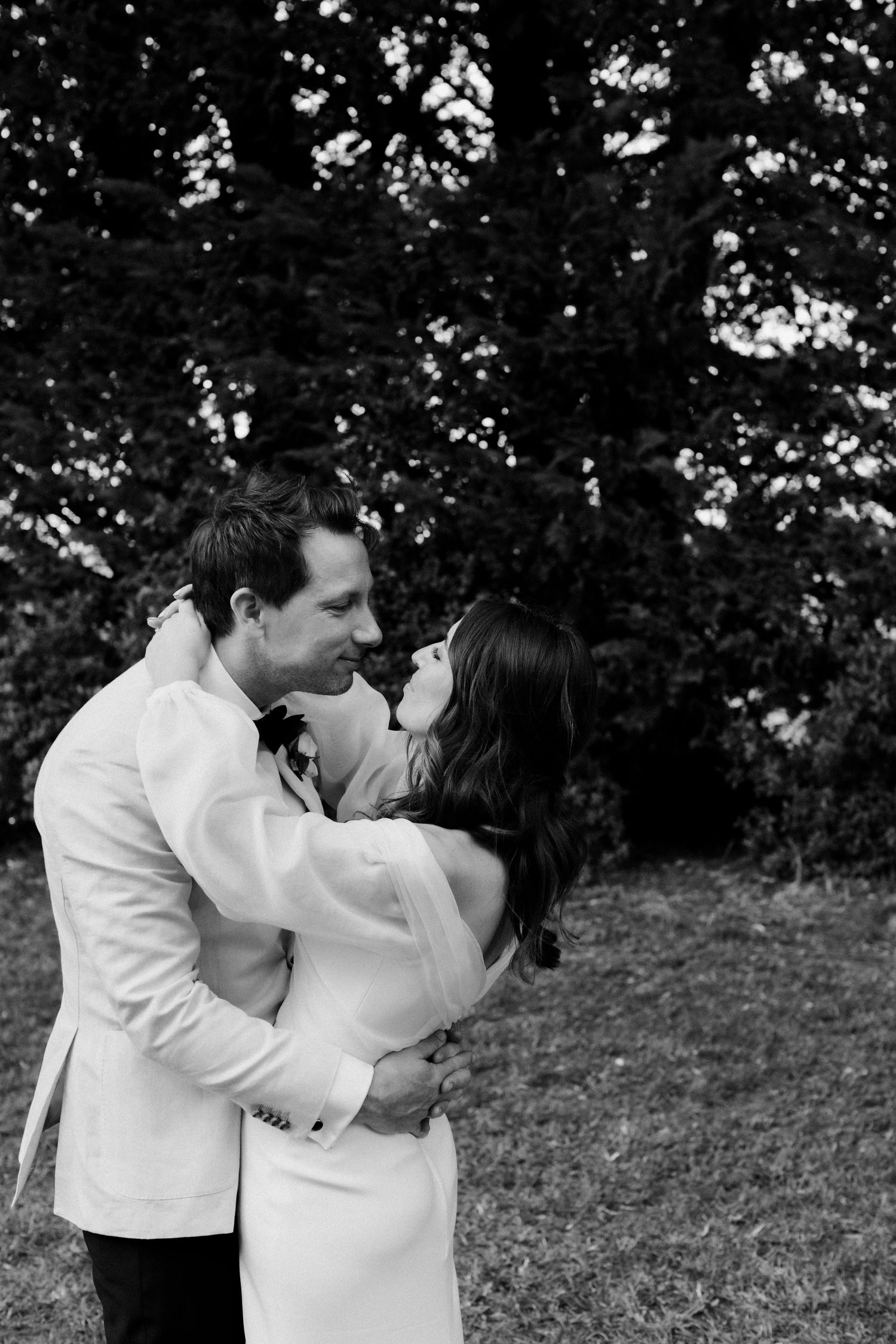 A romantic black and white portrait of a couple embracing outdoors near trees.