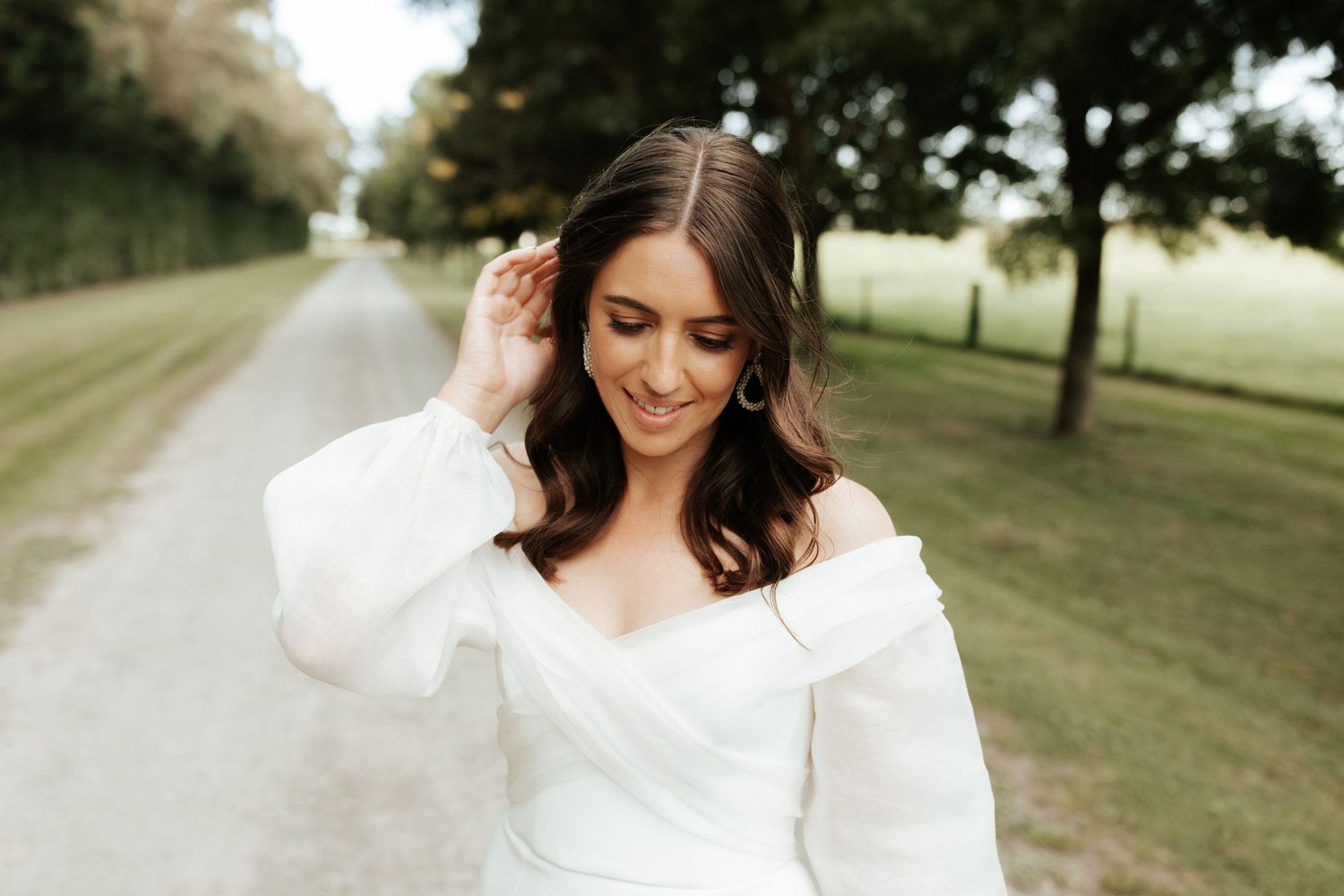 Bride in elegant off-shoulder gown smiles while touching her hair on a countryside path.