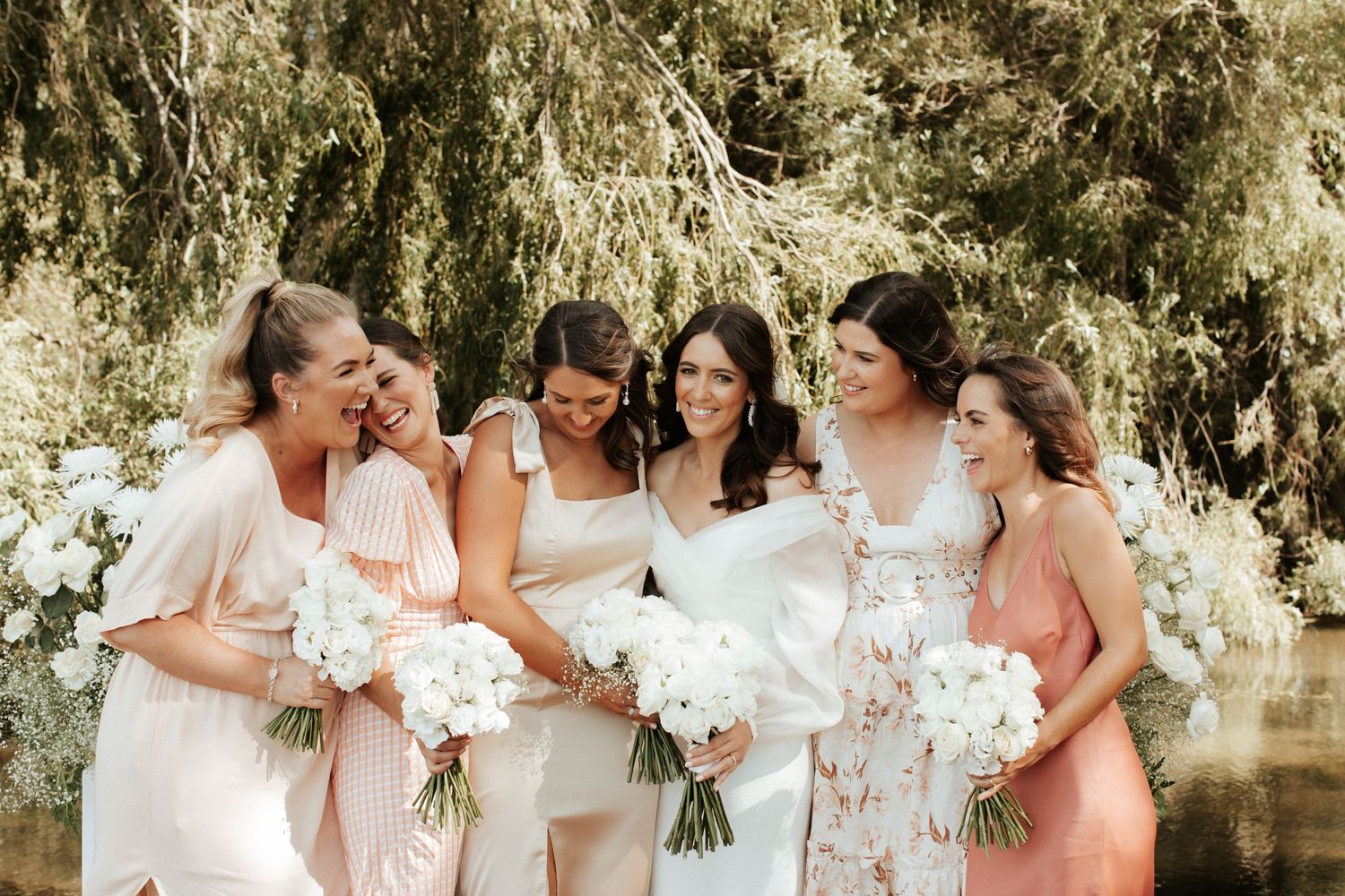 Bridal party in soft pink and white dresses laugh together under a willow tree.