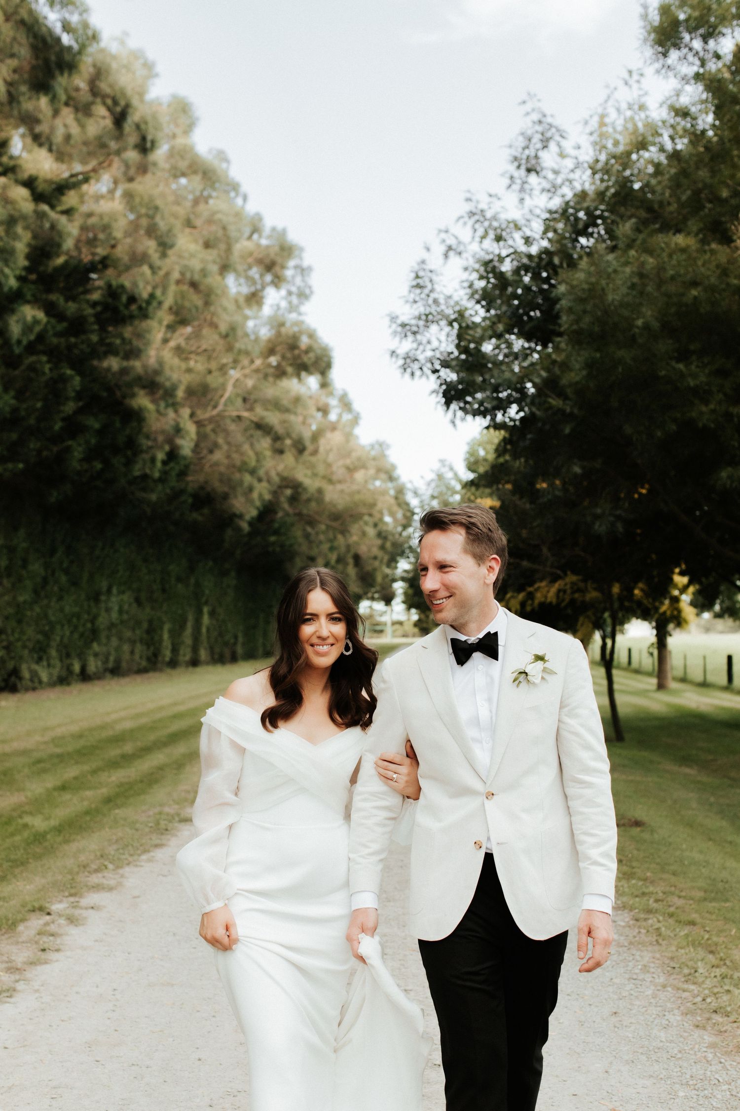 Newlyweds walk arm in arm down a tree-lined path on their wedding day.