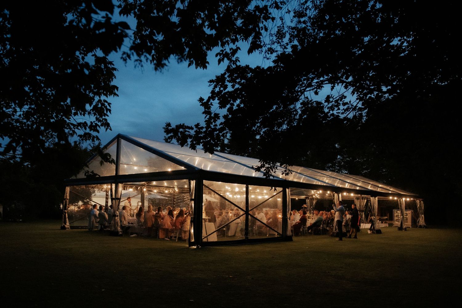 Illuminated clear tent reception venue glowing at dusk with trees in the background.