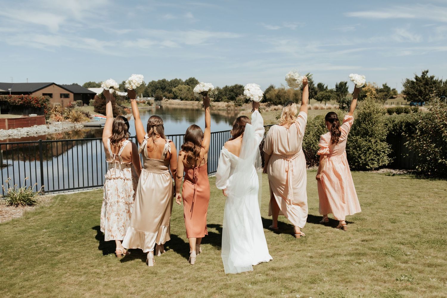 Wedding party celebrates on lawn by waterfront with hands raised joyfully.