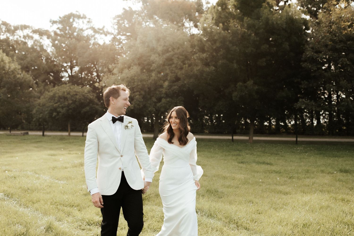 A couple strolls together on their wedding day in a sunlit meadow with trees.