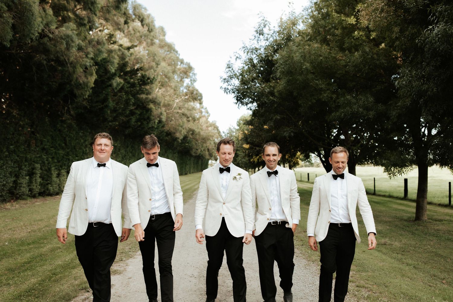 A group of groomsmen in white dinner jackets and black bow ties walk together on a grass path lined with trees.