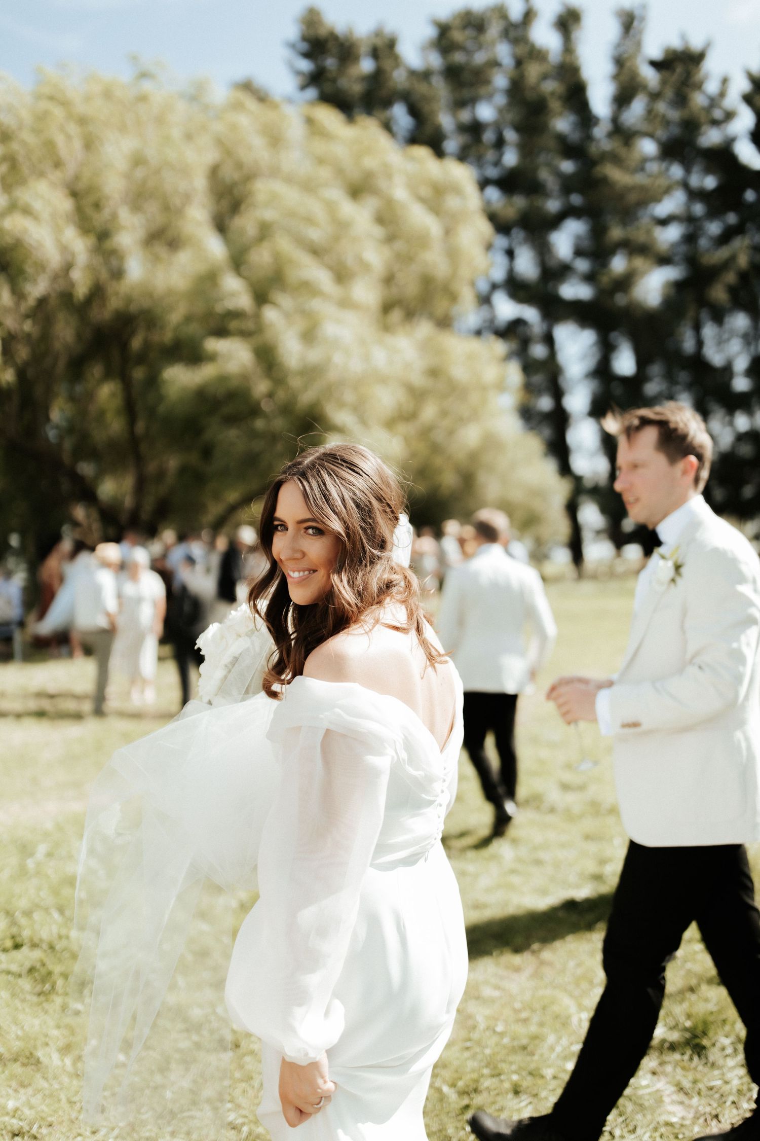 A bride in an elegant white dress walks through a sunny outdoor setting.
