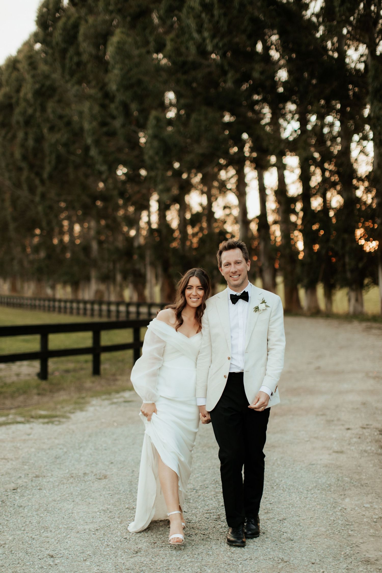 Sunset wedding portrait of couple walking along a fence-lined path.