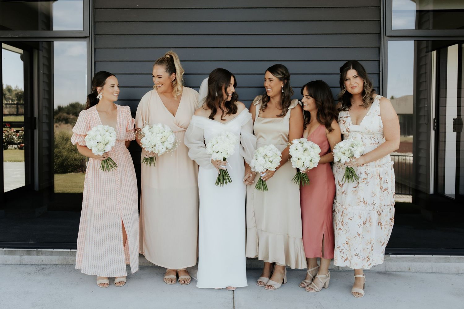 Bridesmaids in soft neutral and floral dresses pose together holding white bouquets.