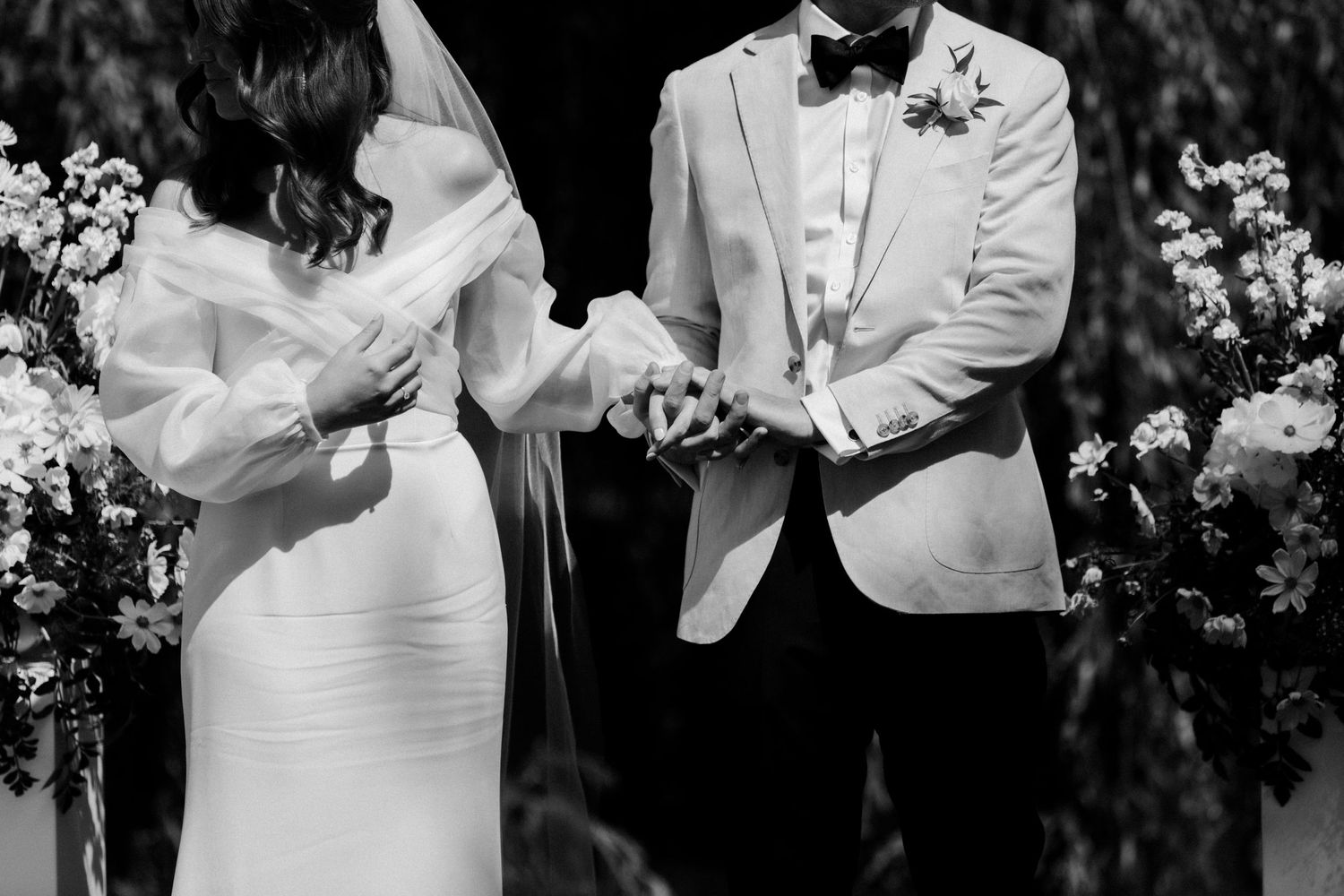 Black and white closeup of couple holding hands during wedding ceremony with floral decor.