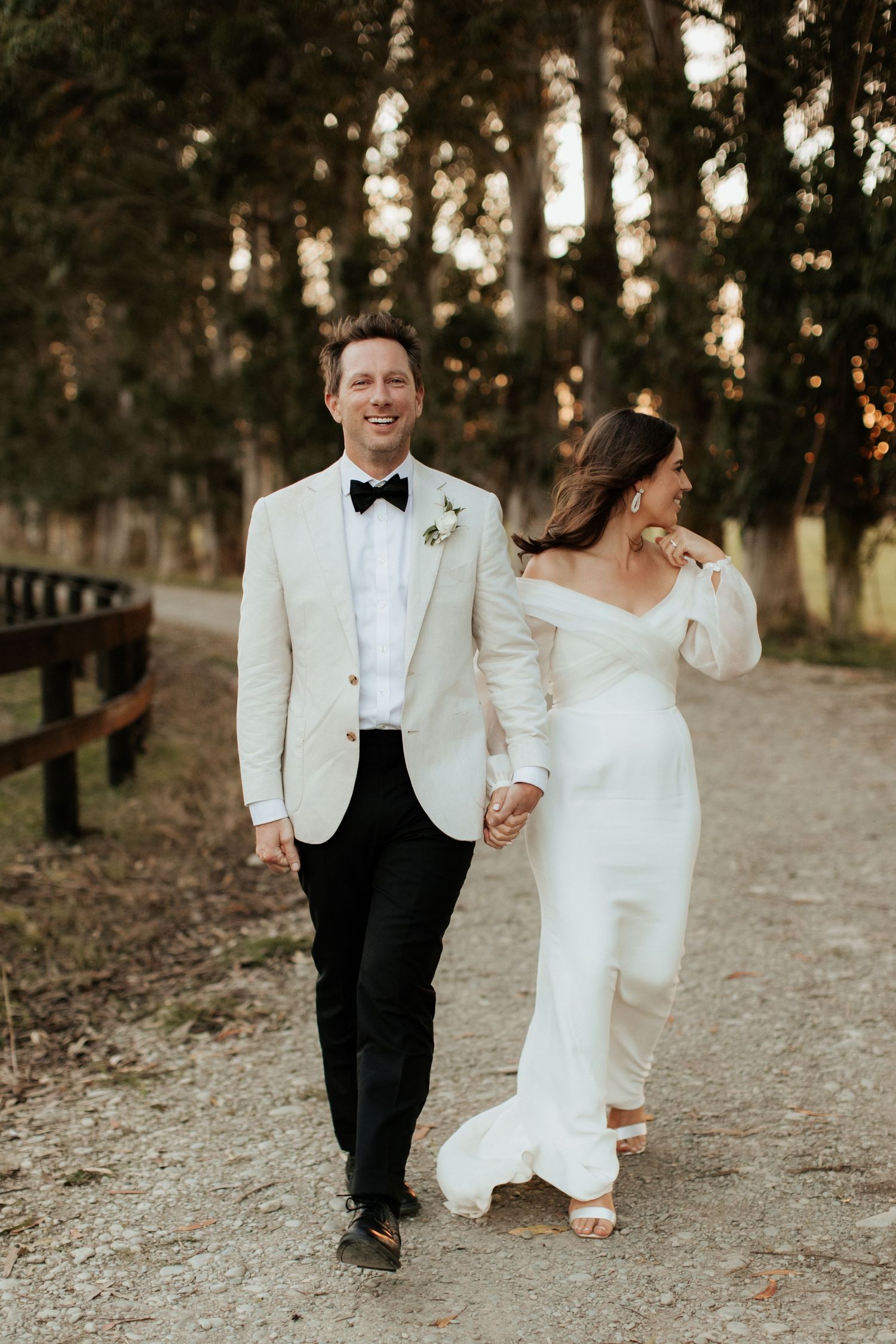 Elegant couple walking hand in hand on a tree-lined path at sunset.