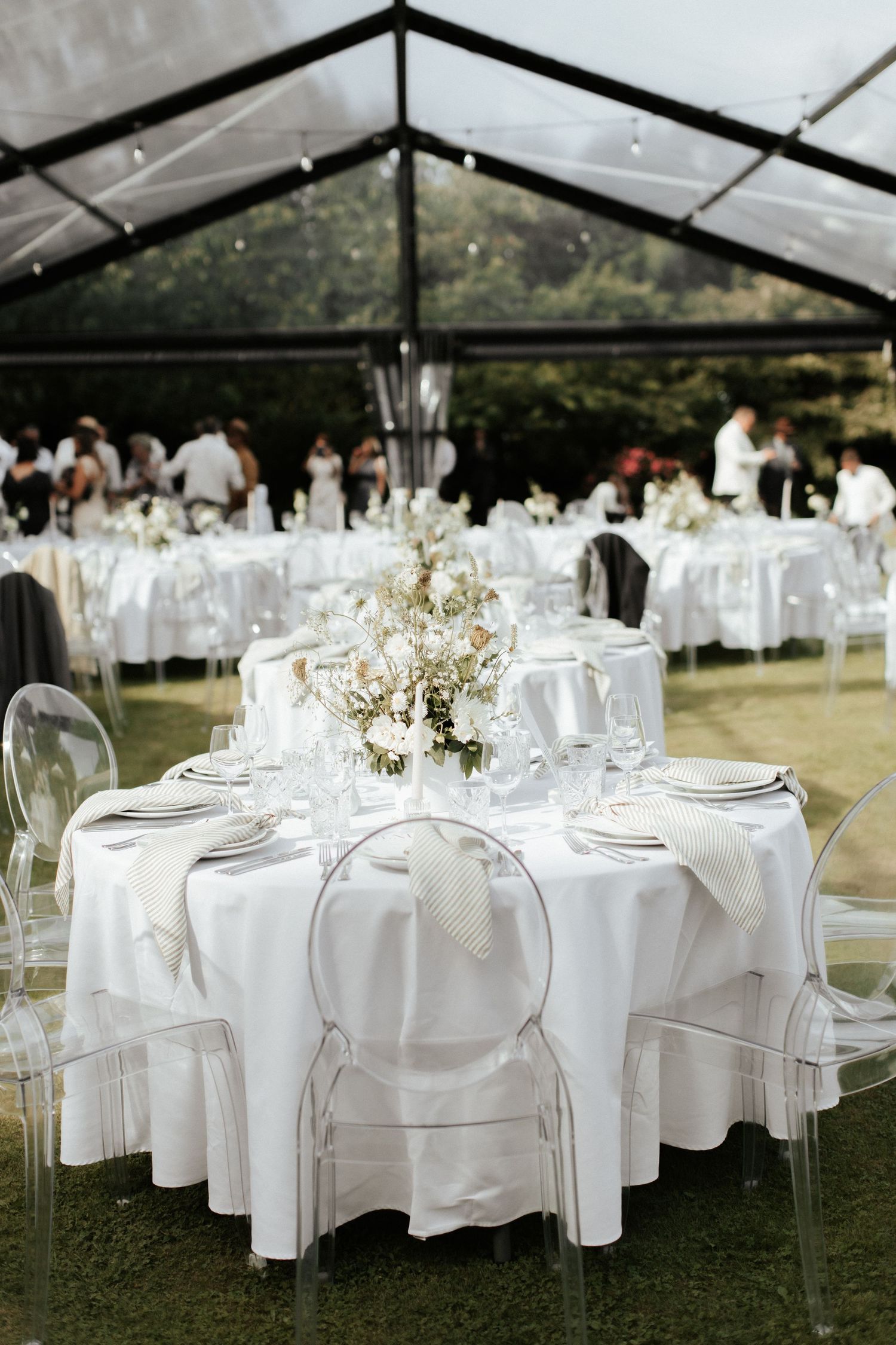 Elegant wedding reception setup with white round tables, clear ghost chairs and delicate floral centerpieces under a tent.