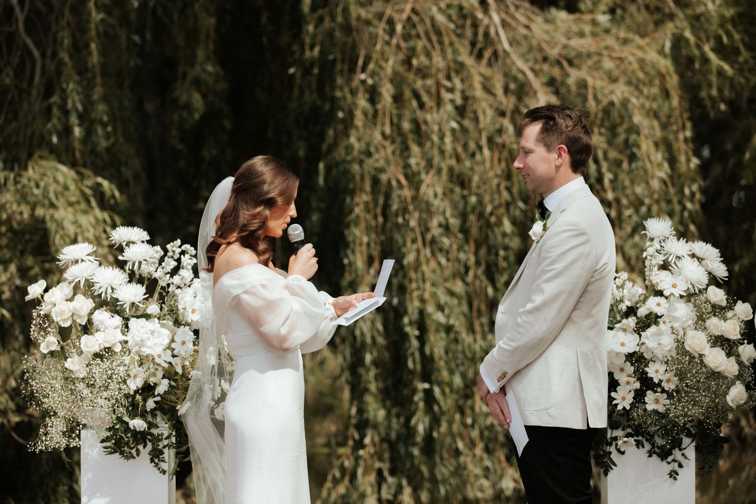 A wedding ceremony under a weeping willow tree with white floral arrangements on pedestals.