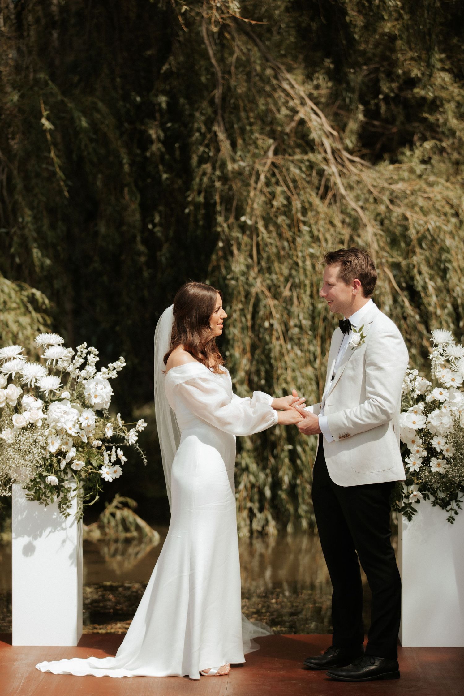 Couple exchanges vows under white floral arrangements with willow trees in background.