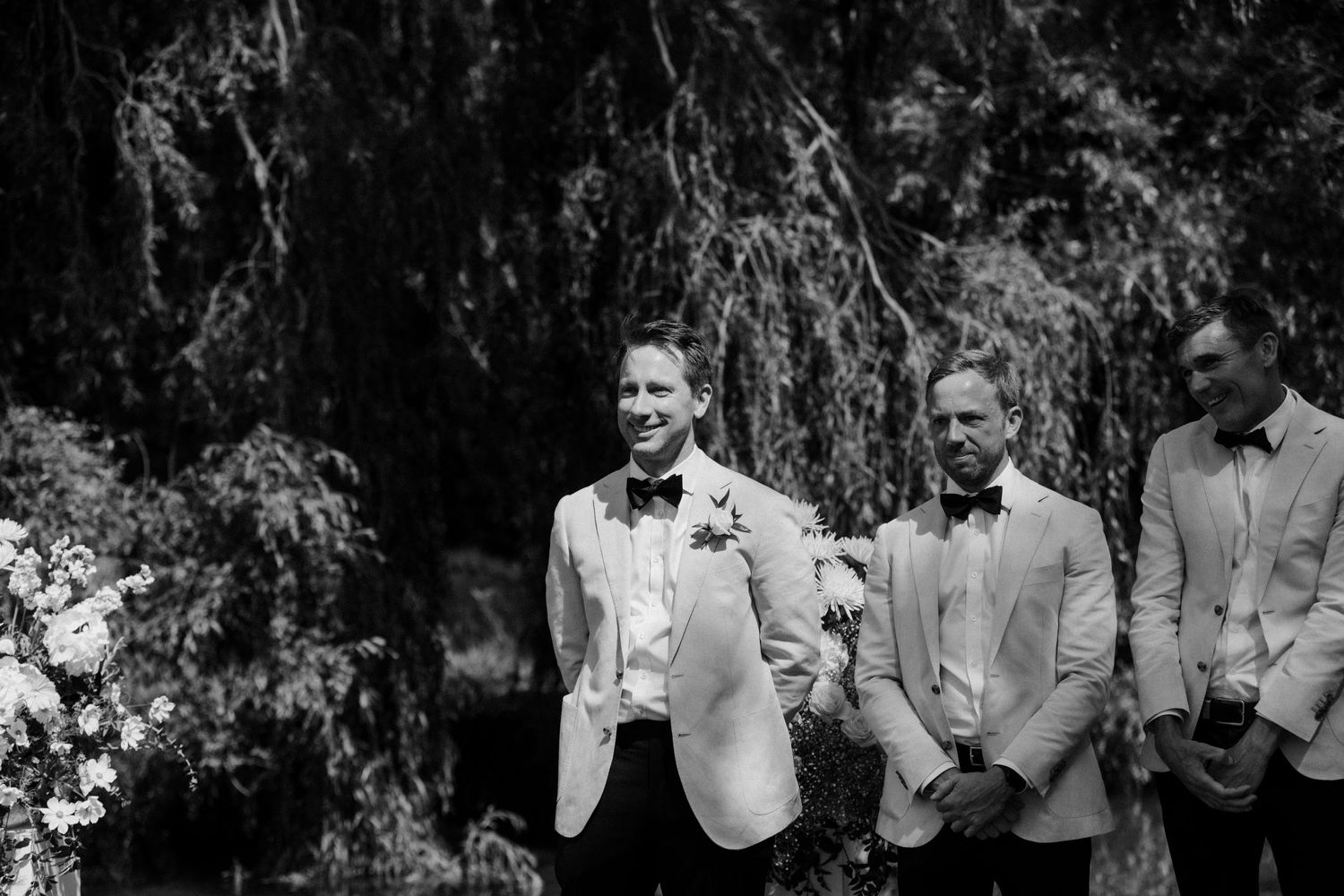Groomsmen in formal attire stand against a backdrop of willow trees.