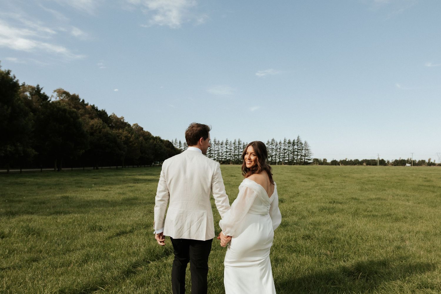Couple in white formal attire walking through a meadow with pine trees in background.
