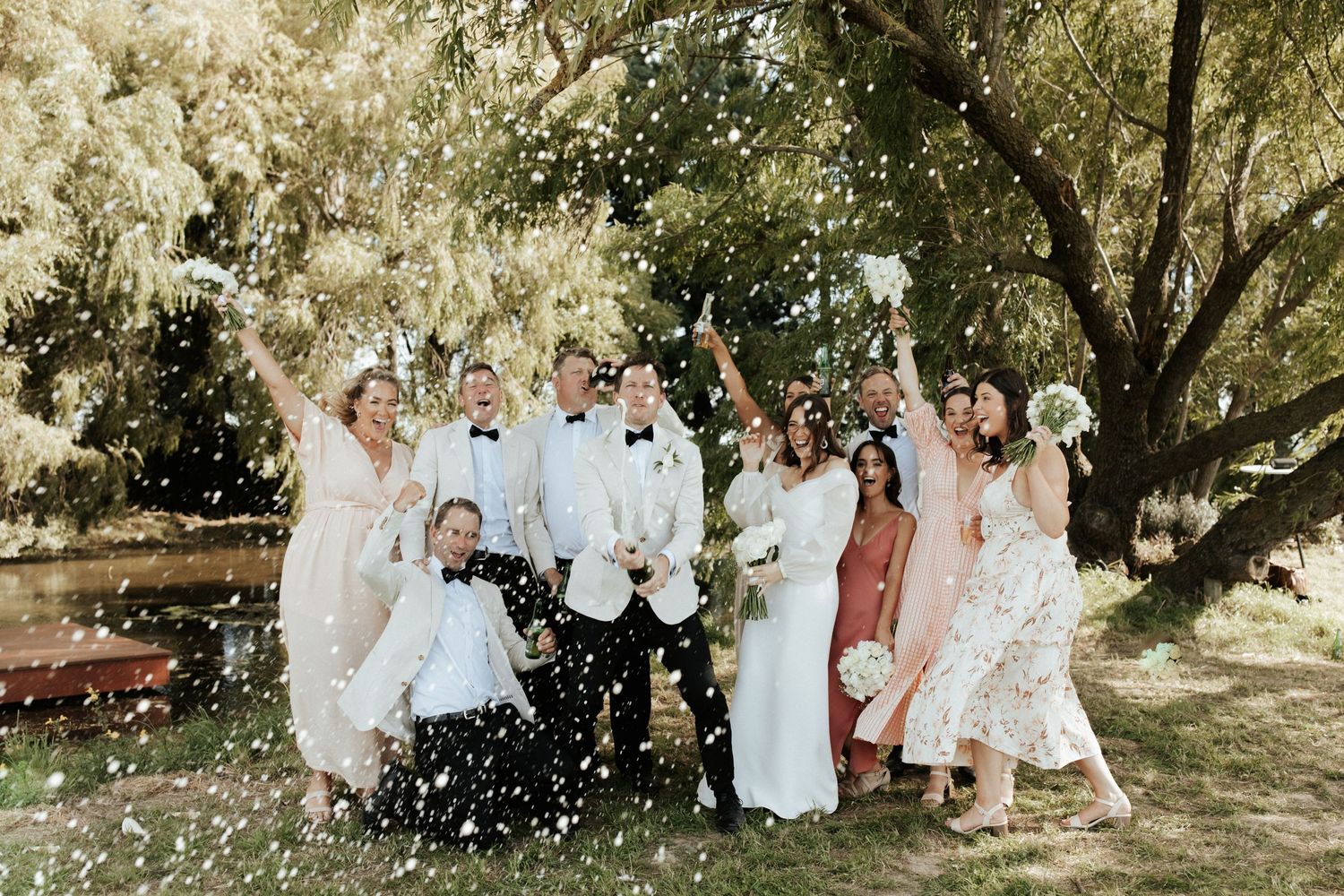 Wedding party celebrates with confetti toss under trees at outdoor celebration.