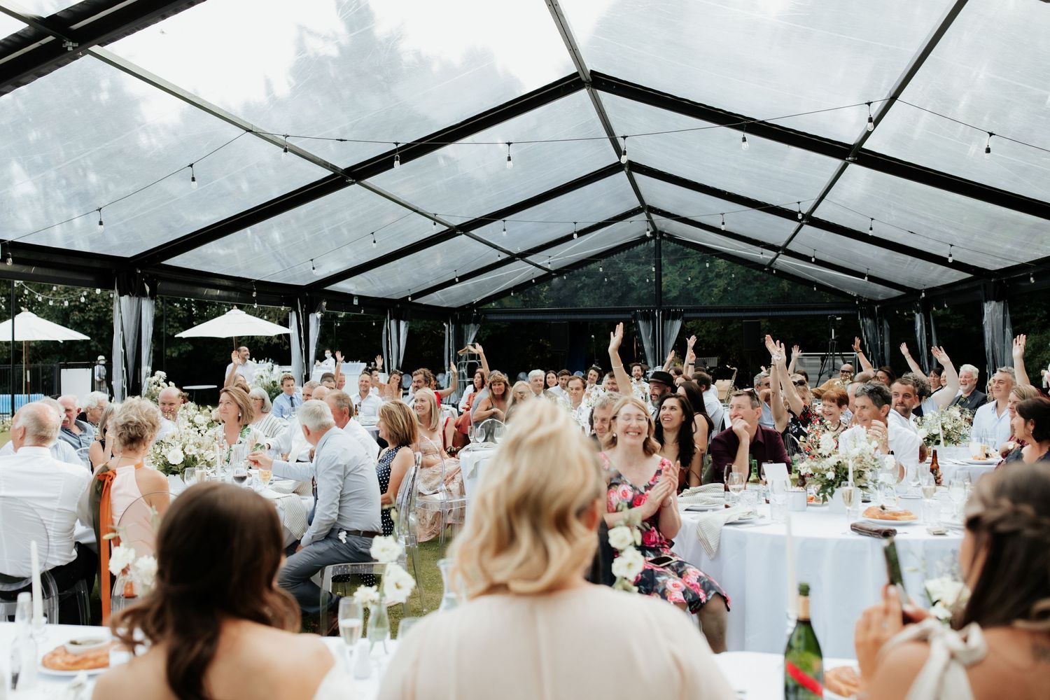 Wedding reception guests raise their hands in celebration under a clear-top tent.