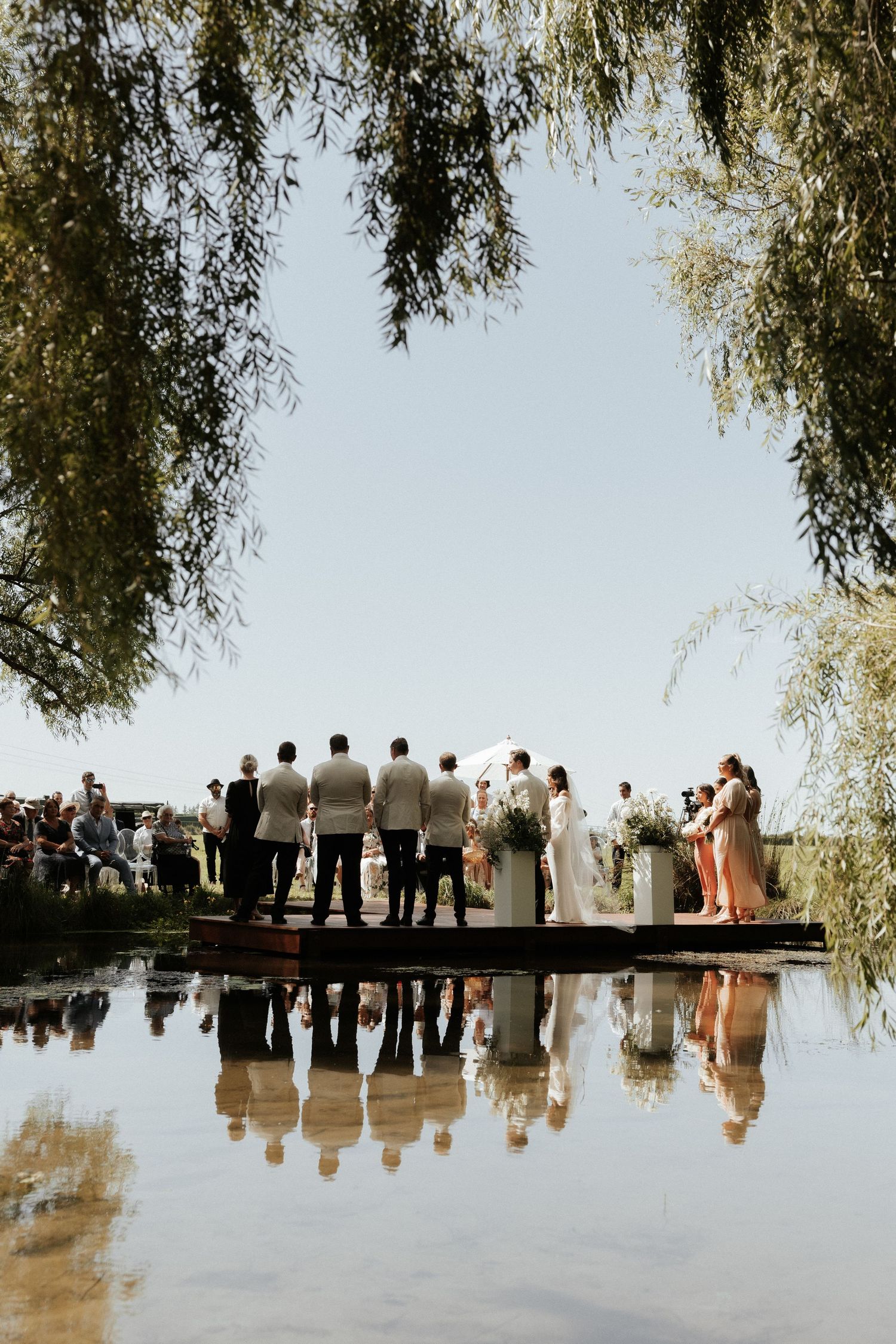 Wedding ceremony reflection in water with willow trees framing the scene.