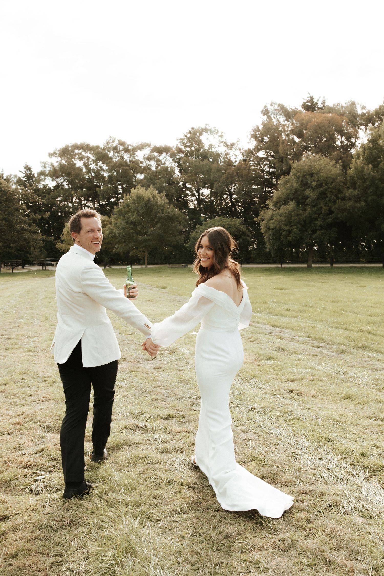 Couple in white formal attire dancing and smiling in a sunlit field.