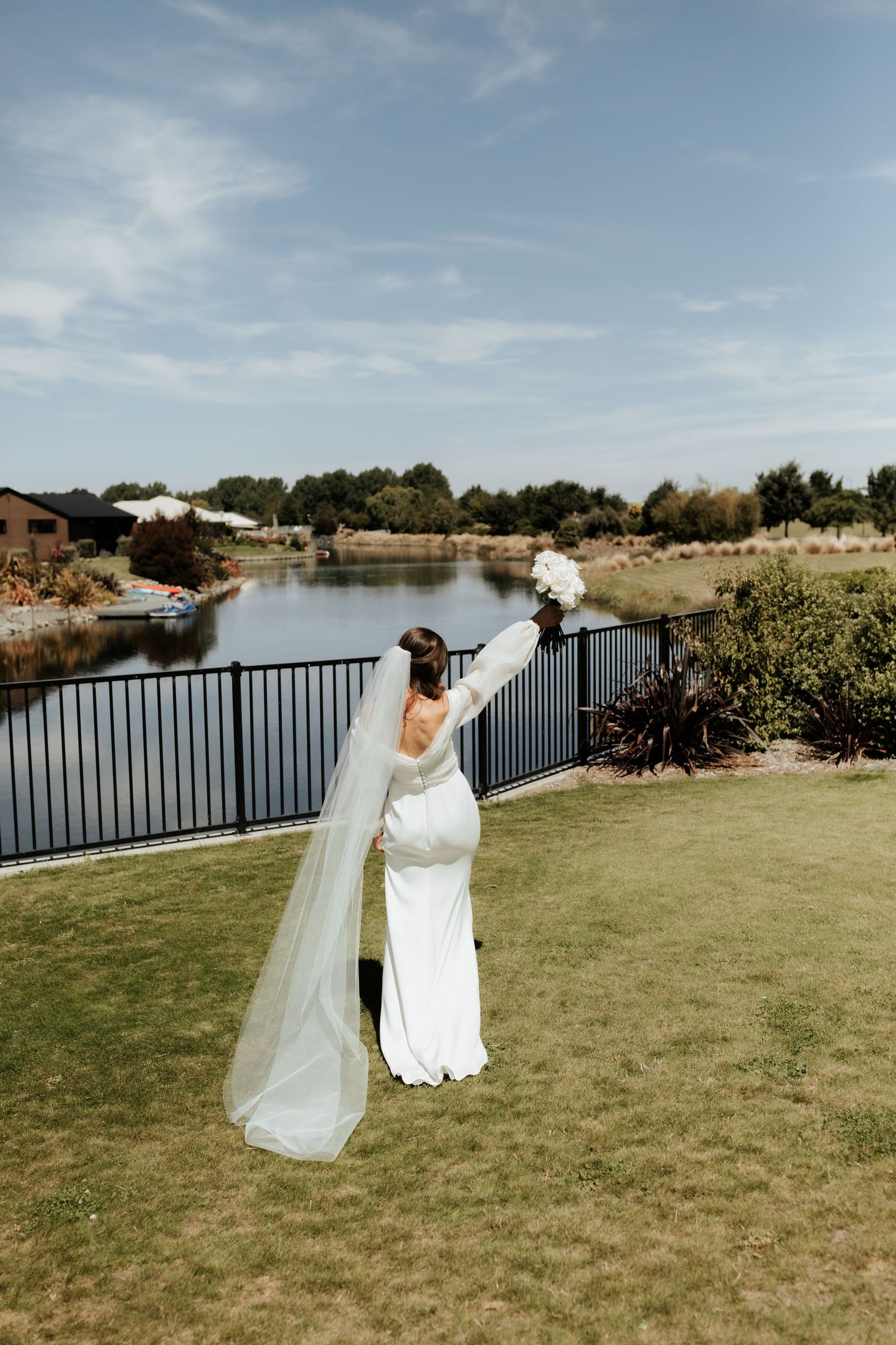 Bride in fitted white gown and long veil stands by lakeside on manicured lawn.