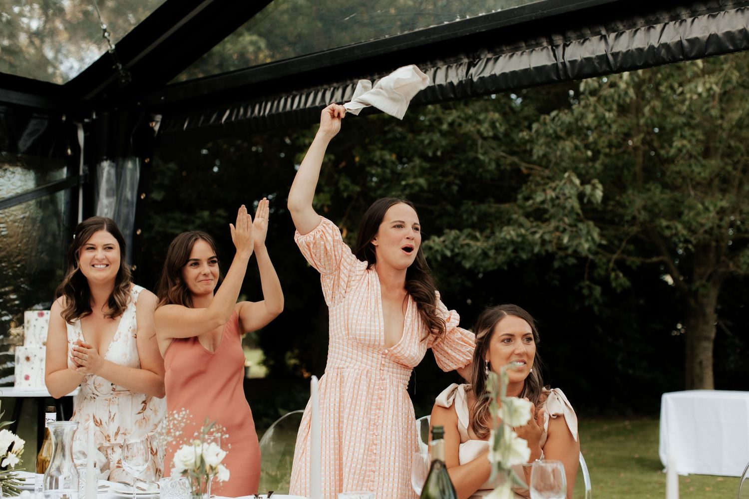 Wedding guests celebrate enthusiastically during reception under clear tent.