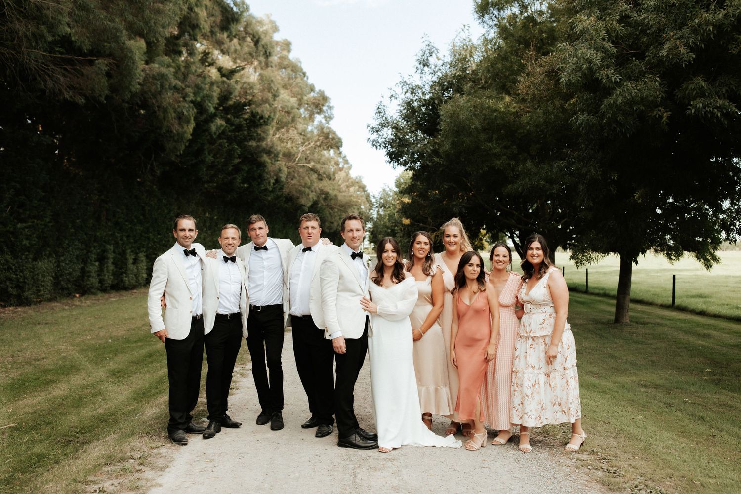 A wedding party poses together outdoors with bridesmaids in pink dresses and groomsmen in white jackets.