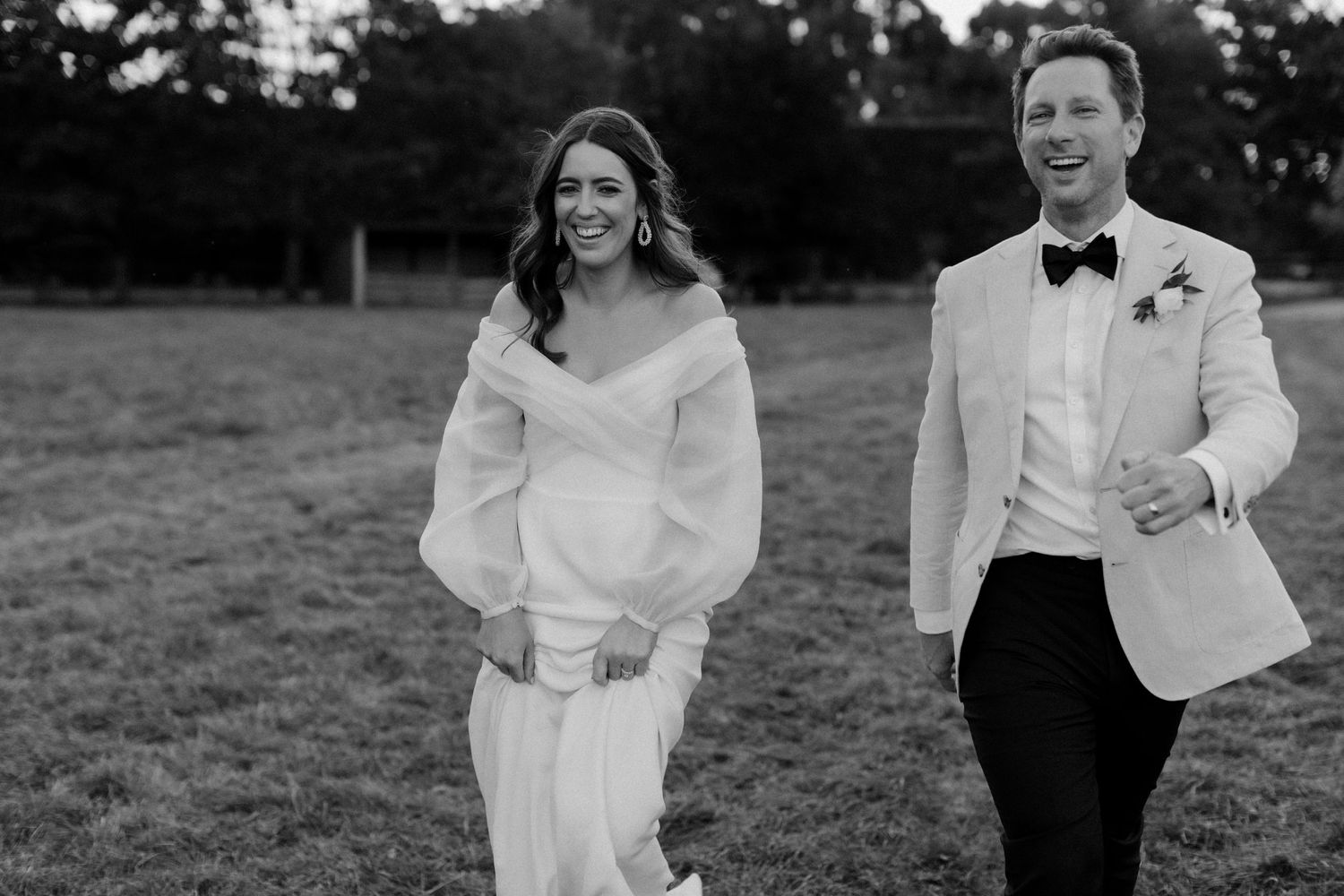 A couple in wedding attire walks together and laughs in a black and white outdoor photo.
