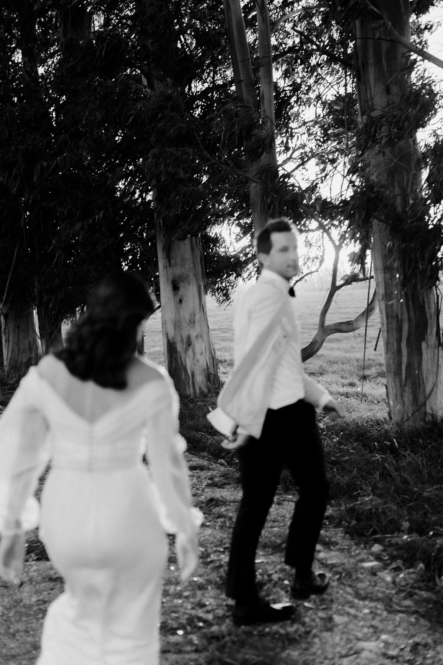 Couple in wedding attire dancing playfully among eucalyptus trees.