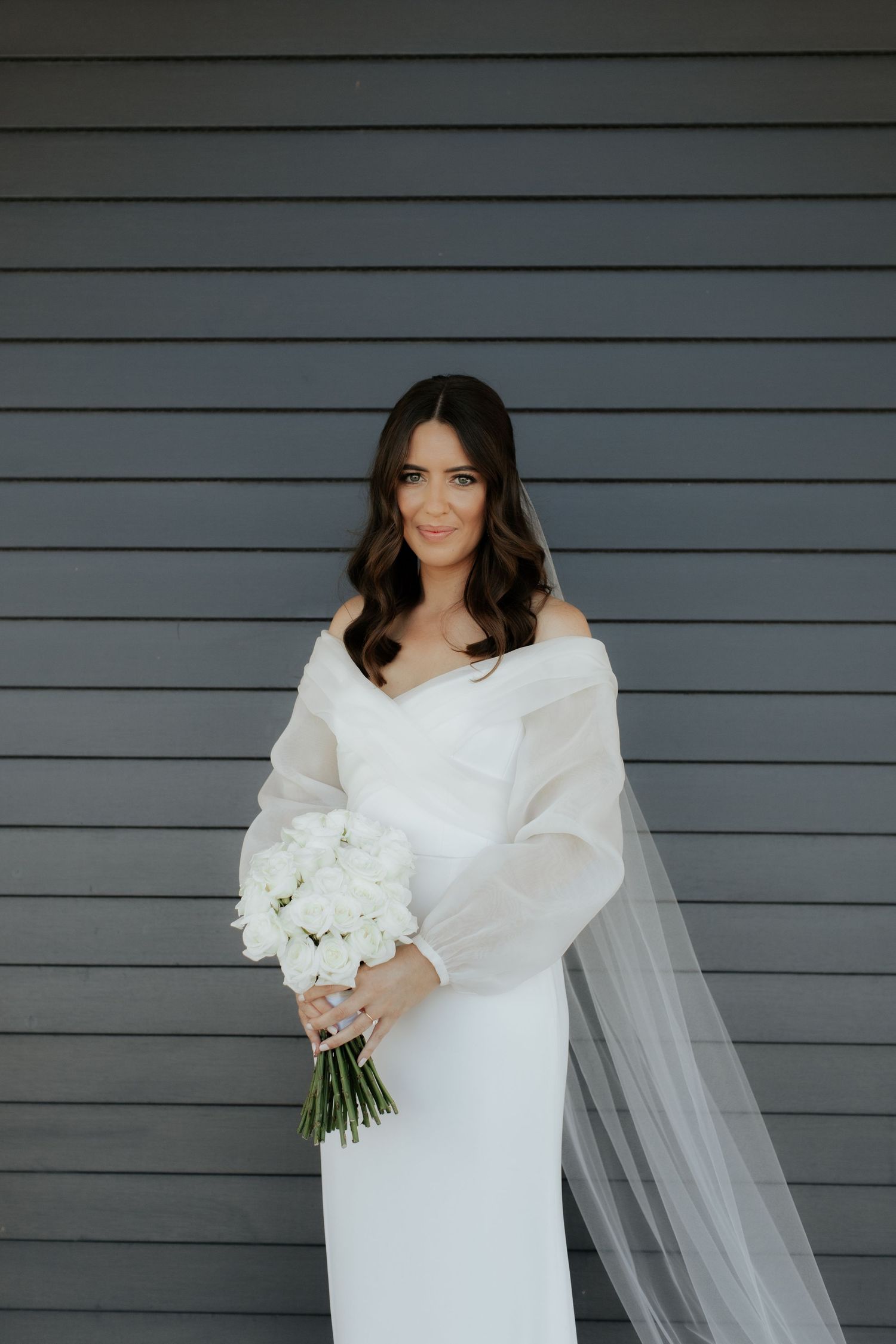 Bride poses with white bouquet against modern gray siding.