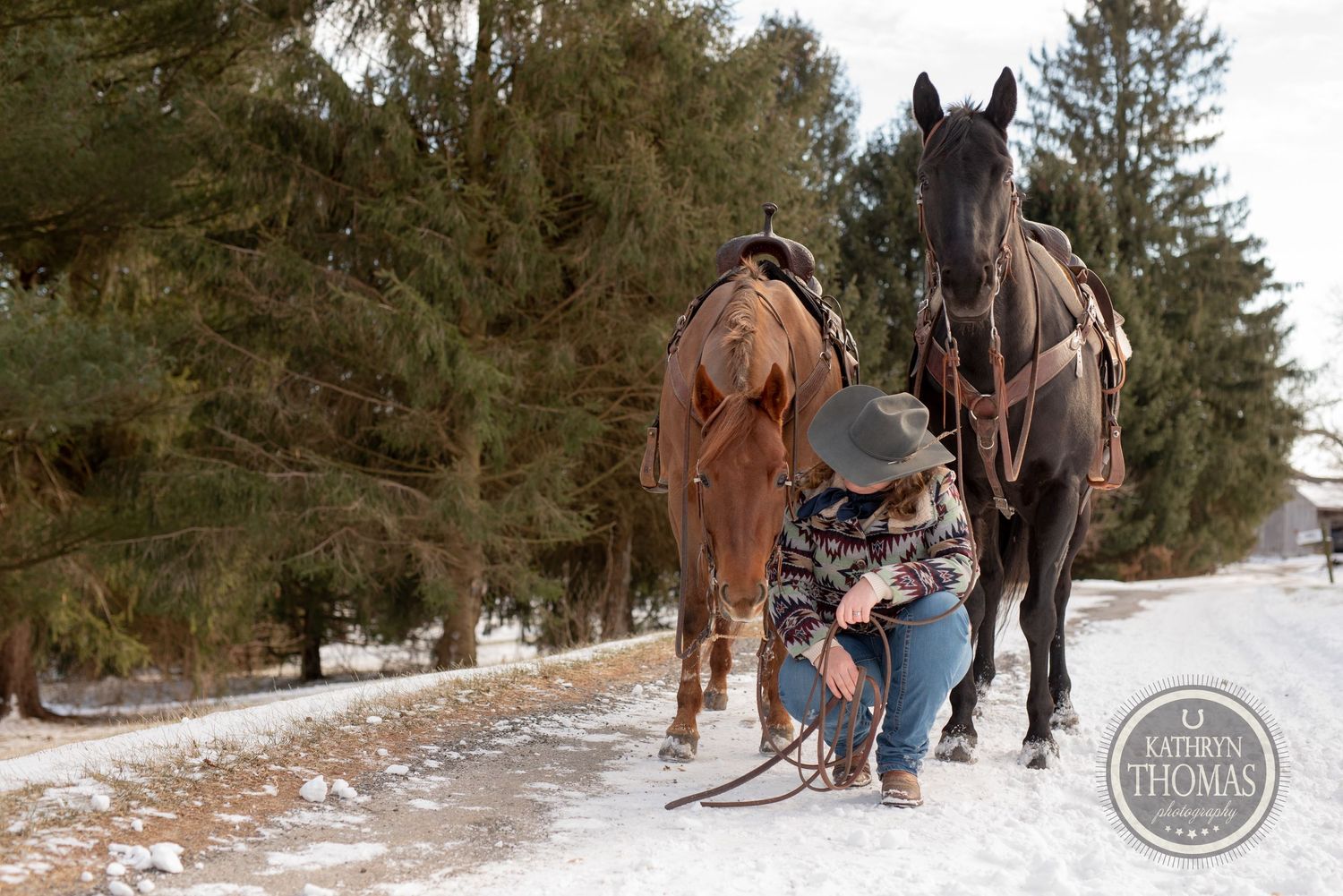 A person adjusts tack on two saddled horses on a snow-covered path.