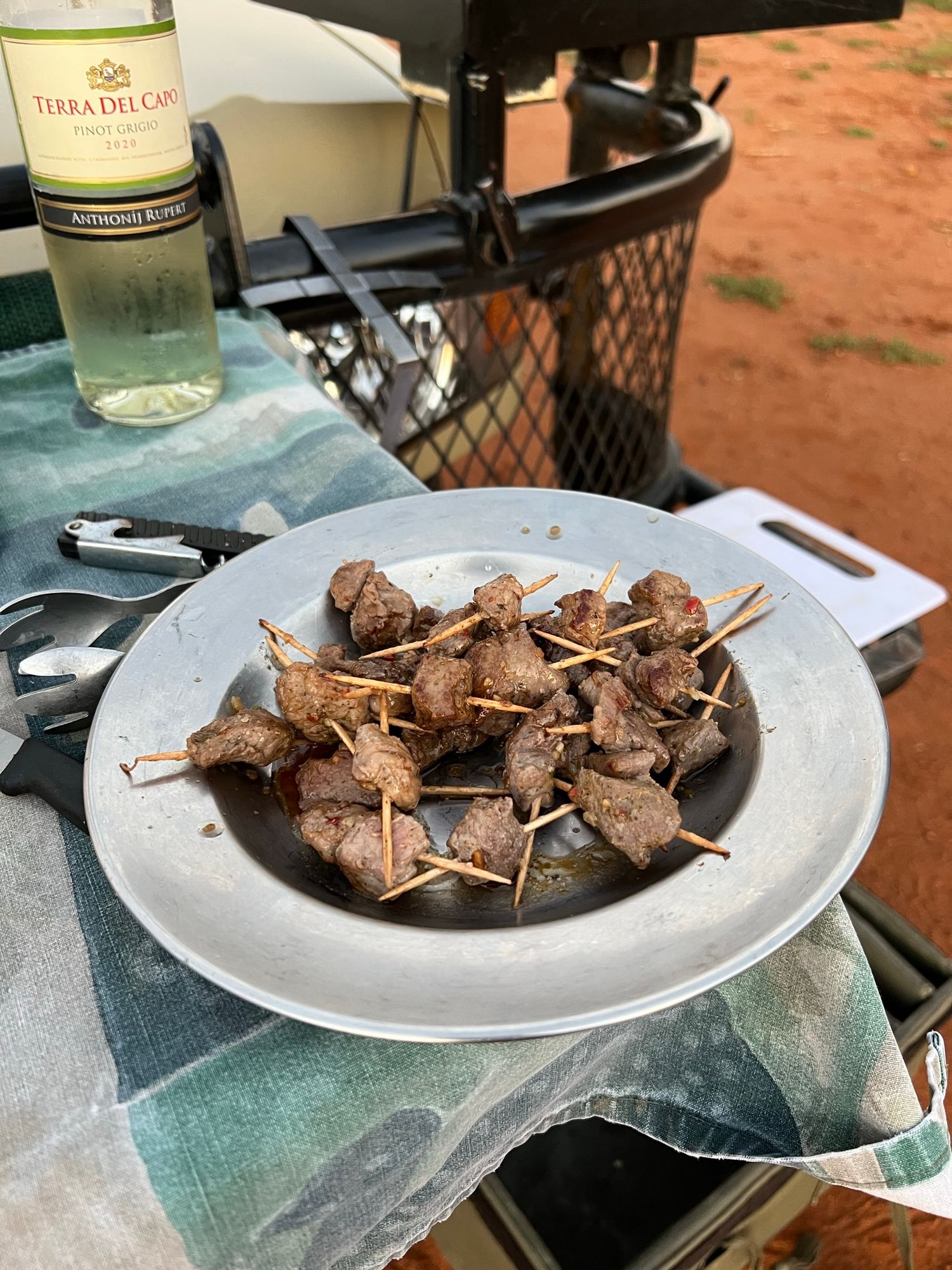 Grilled meat skewers served on white plates with wine bottles in outdoor setting.