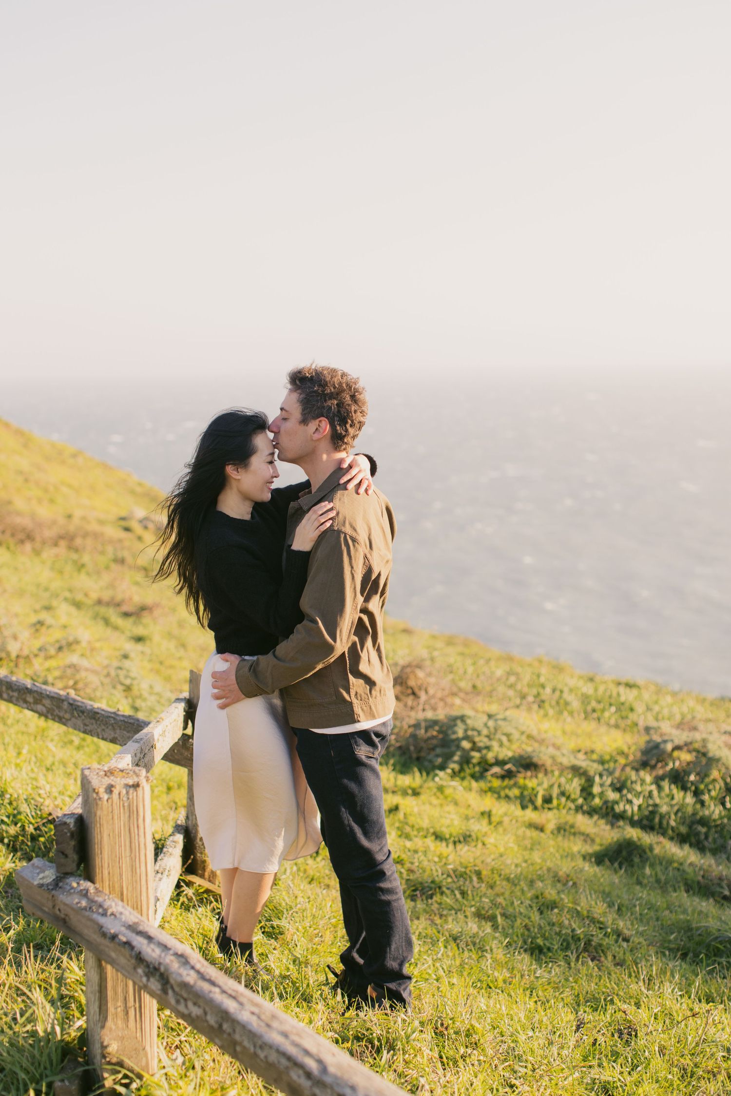 A romantic moment shared between a couple on a coastal hillside with wooden fencing.