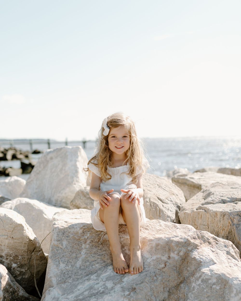 Little girl in light dress sits happily on coastal rocks by ocean on sunny day.