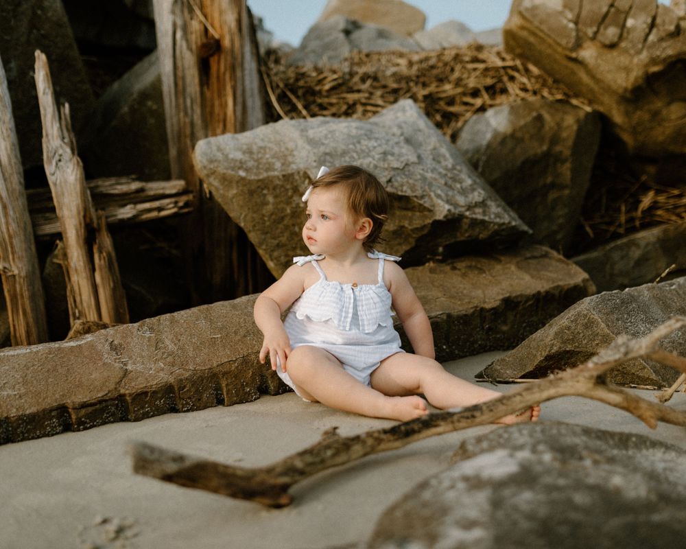 Young child in white clothing sitting on driftwood among large rocks at sunset.