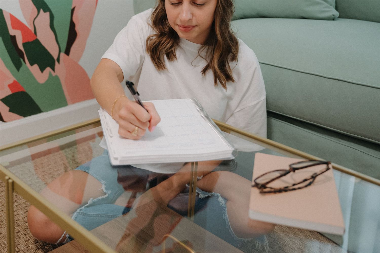 Someone writing in a notebook on a glass coffee table with a modern interior setting.