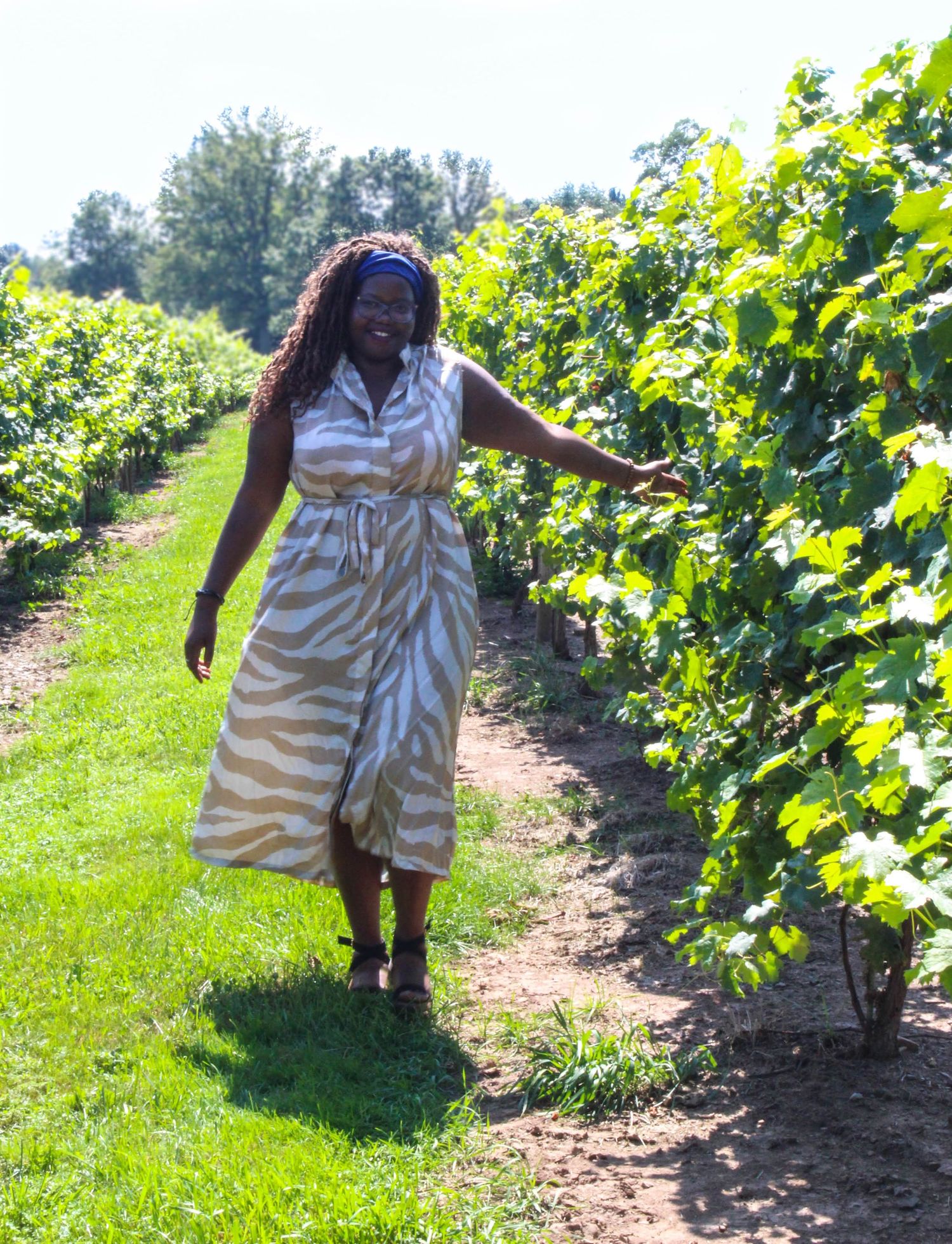 A person explores a sunny vineyard wearing a white dress and blue headband.