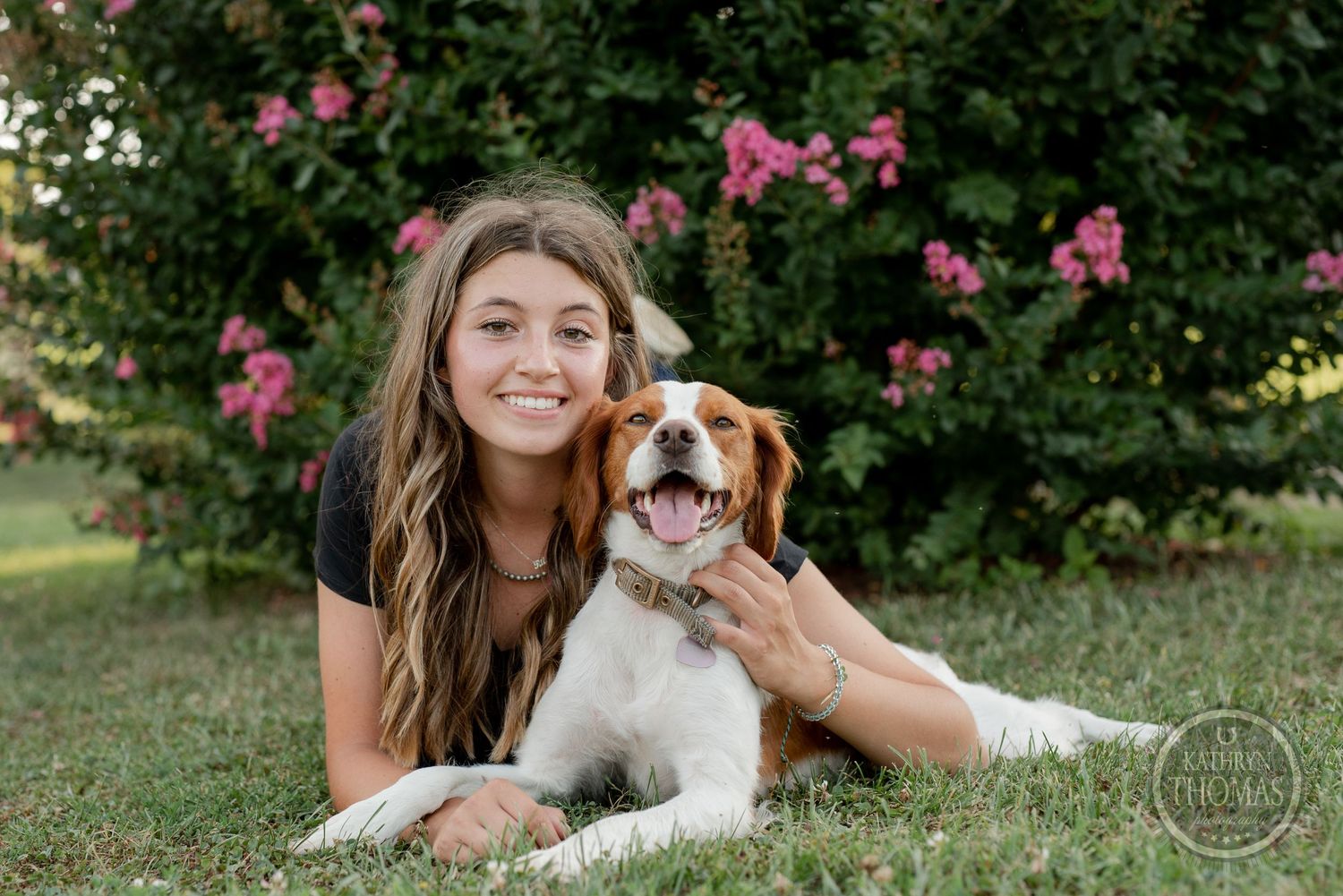 Outdoor portrait with a happy brown and white dog lying in grass with pink flowers behind.