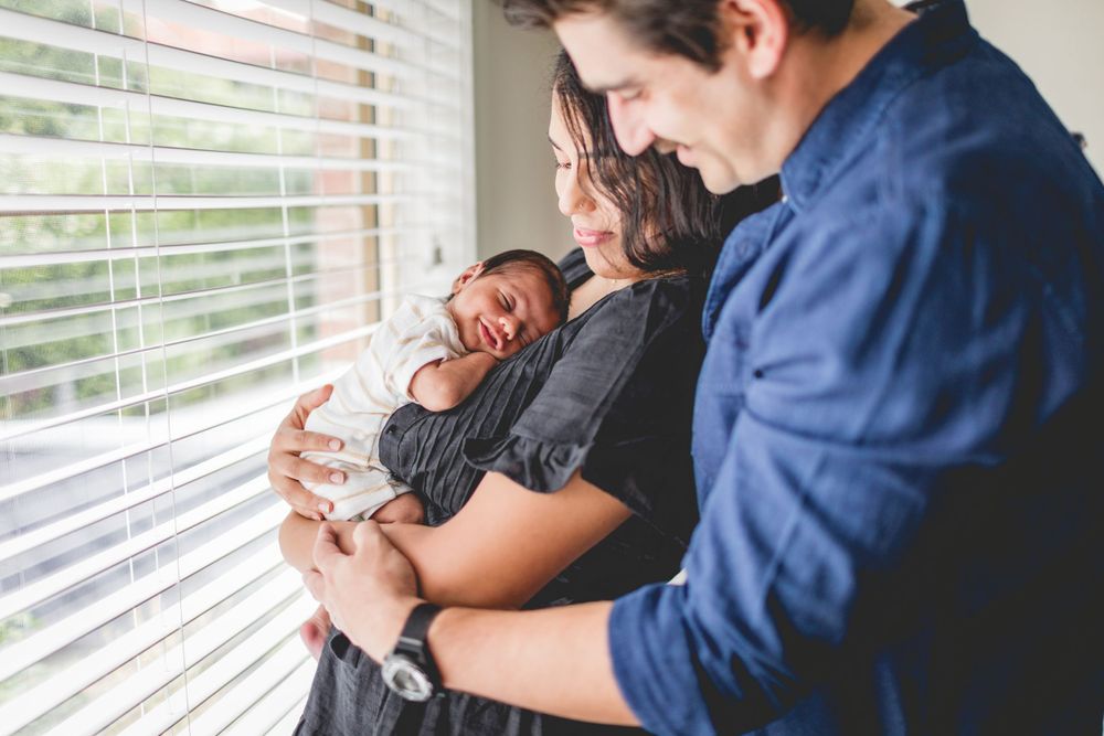 A couple holding their newborn baby next to a sunny window with venetian blinds.