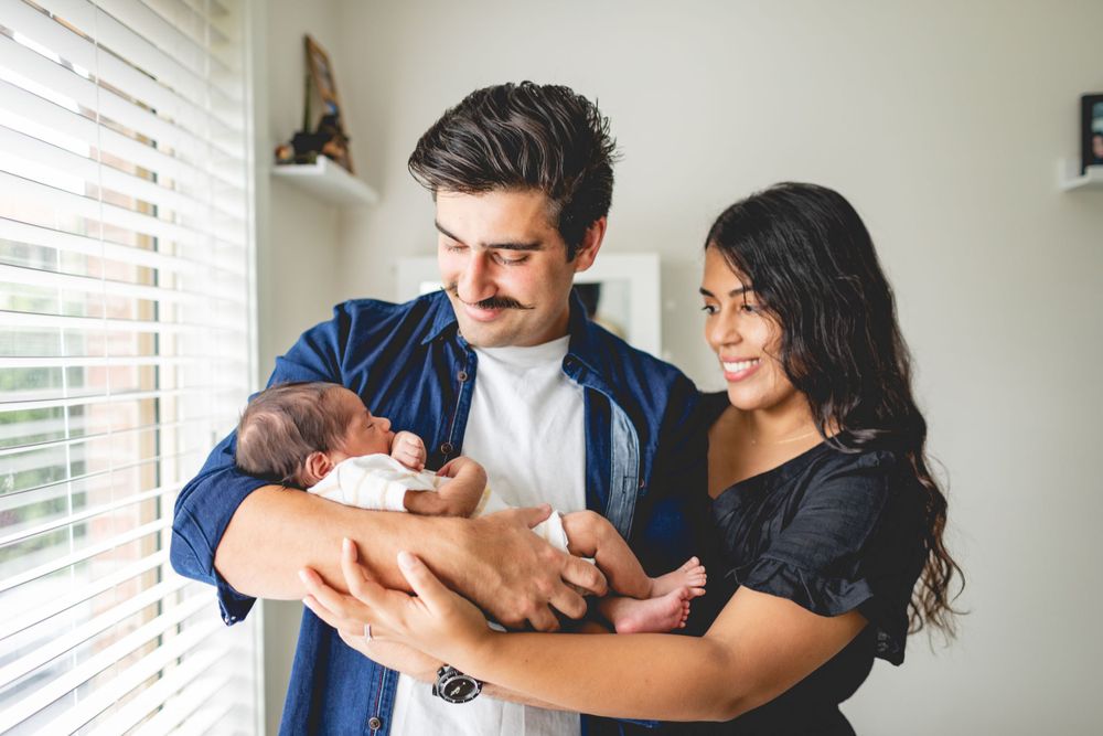 Parents in casual attire lovingly holding their newborn baby near a bright window.