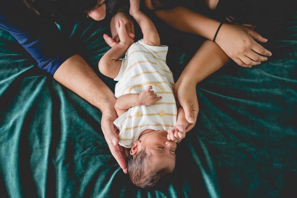 A baby in white clothing lies on a teal blanket while being supported by caring hands.