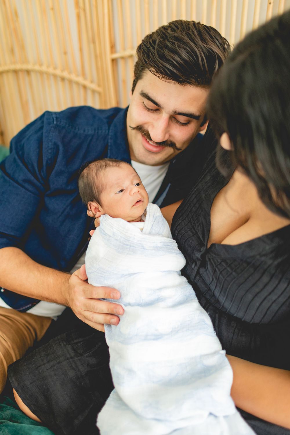 Sweet family moment with swaddled newborn baby against rattan headboard.