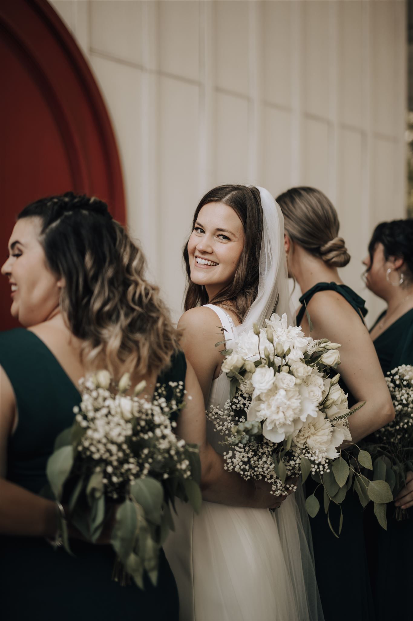 Wedding party members smile while holding elegant white floral bouquets with greenery.