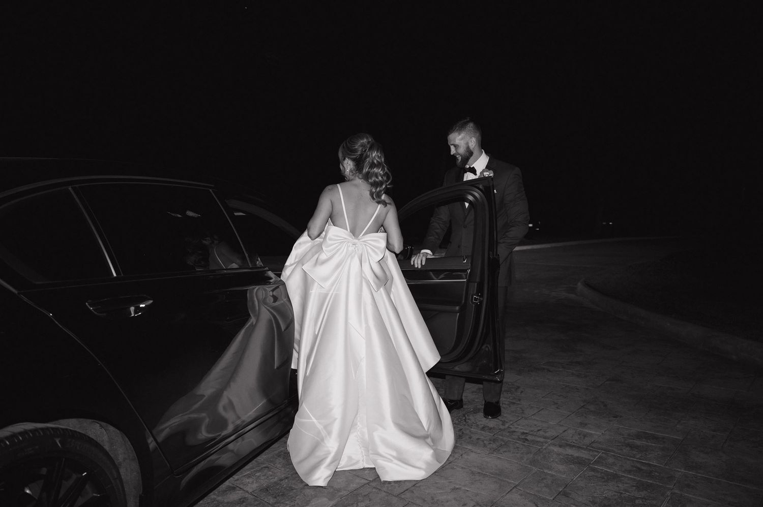 Black and white photo of a couple on their wedding night standing by a car, with the bride wearing a full satin gown.