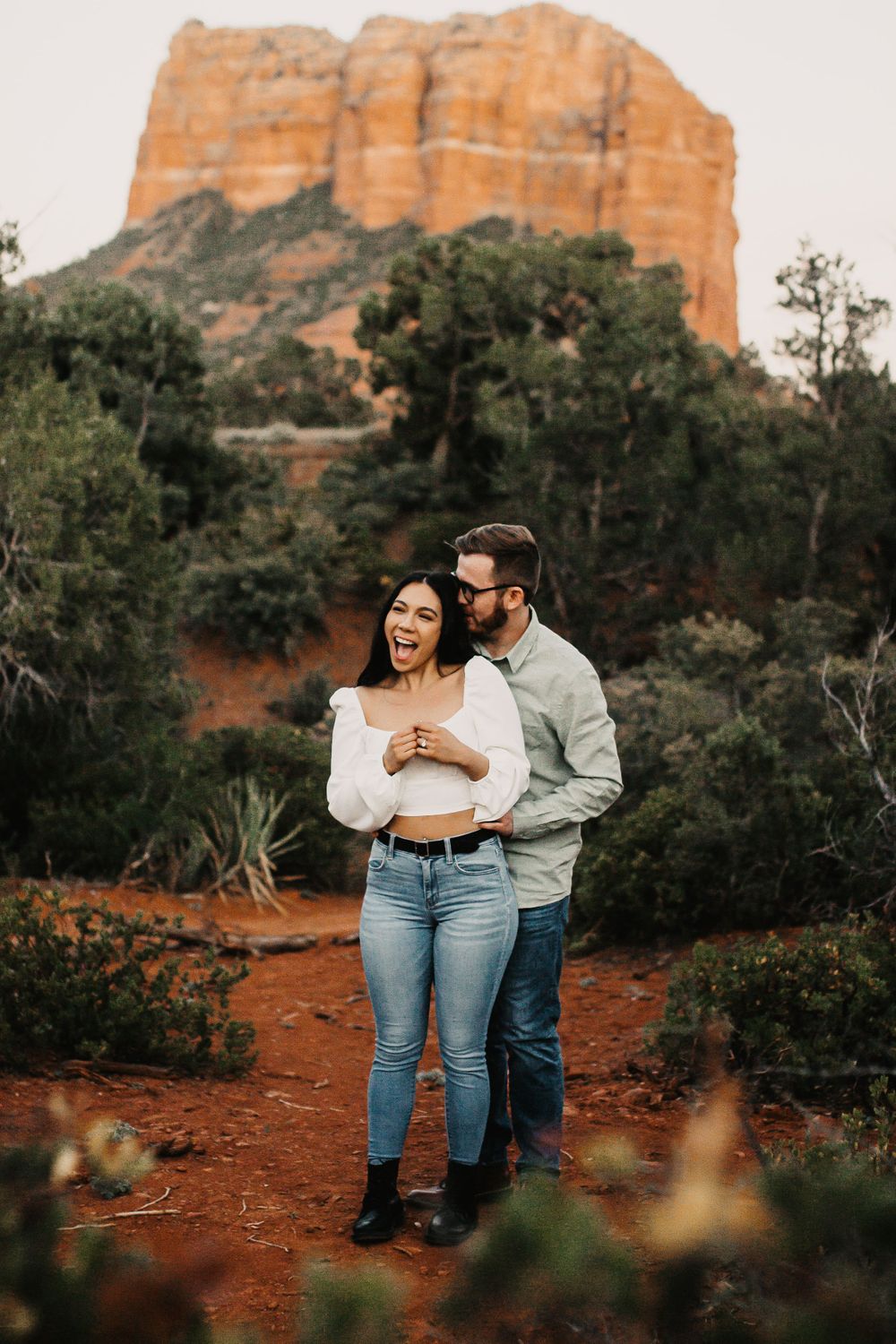 Couple laughing together on red desert soil with towering cliffs behind them.