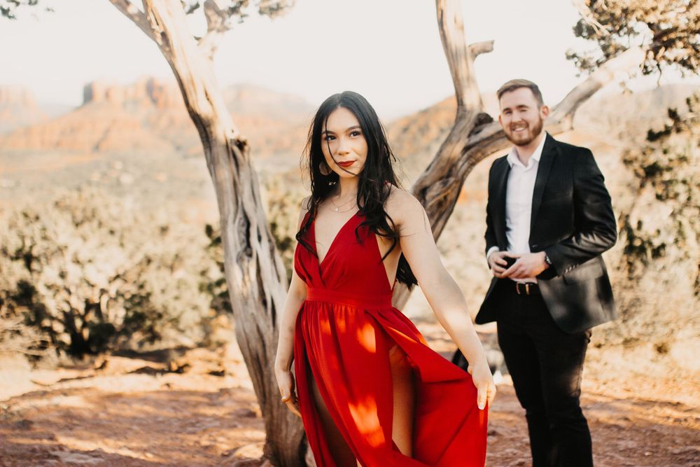 A couple poses among desert trees, with her in a flowing red dress and him in formal attire.