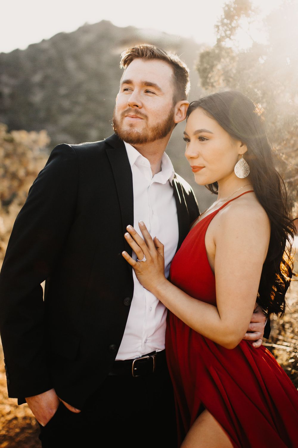 Romantic couple embraces during golden hour photoshoot in nature, with woman in flowing red dress.